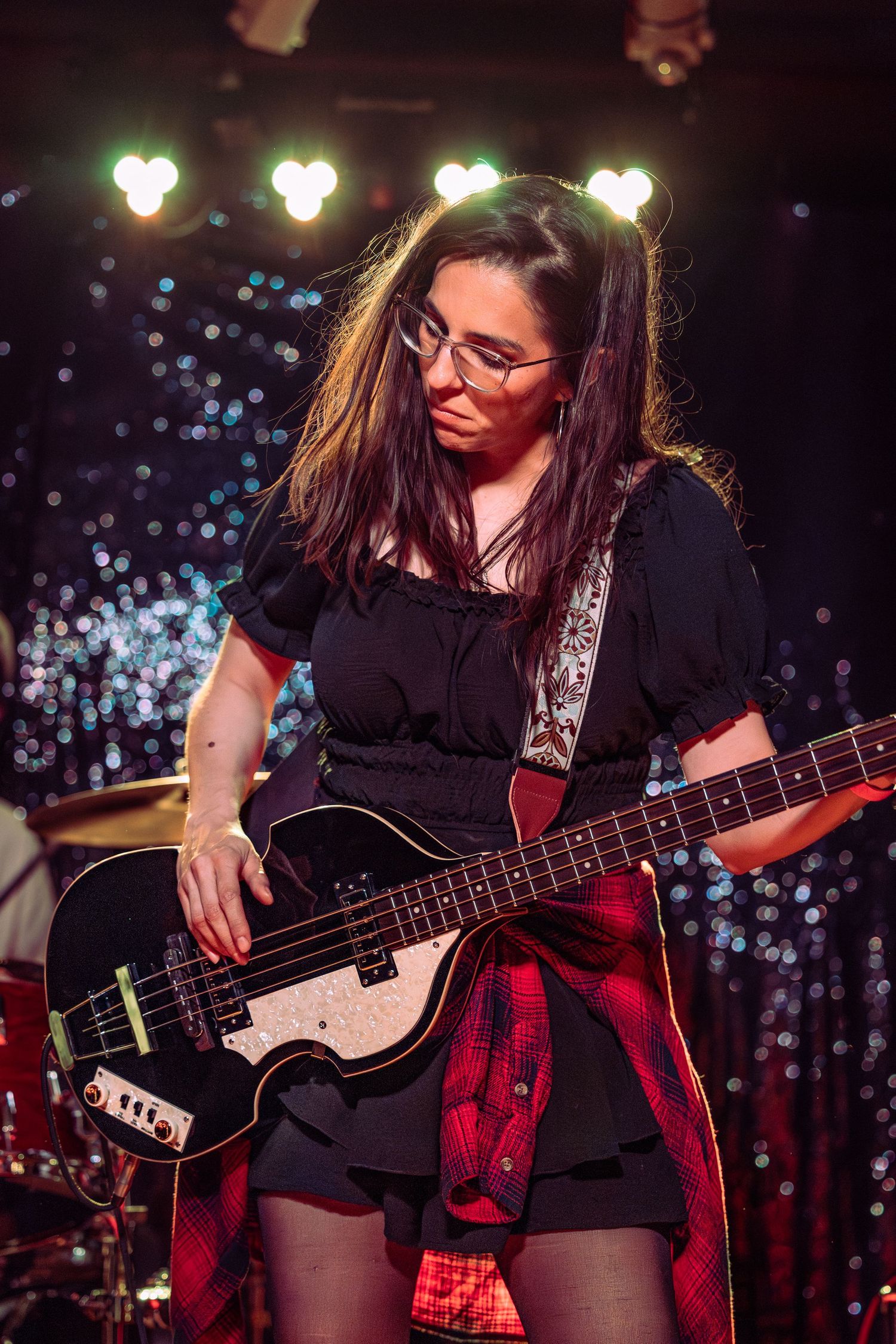 Female bassist with glasses in black dress playing dark Hofner bass guitar under sparkling stage lights.
