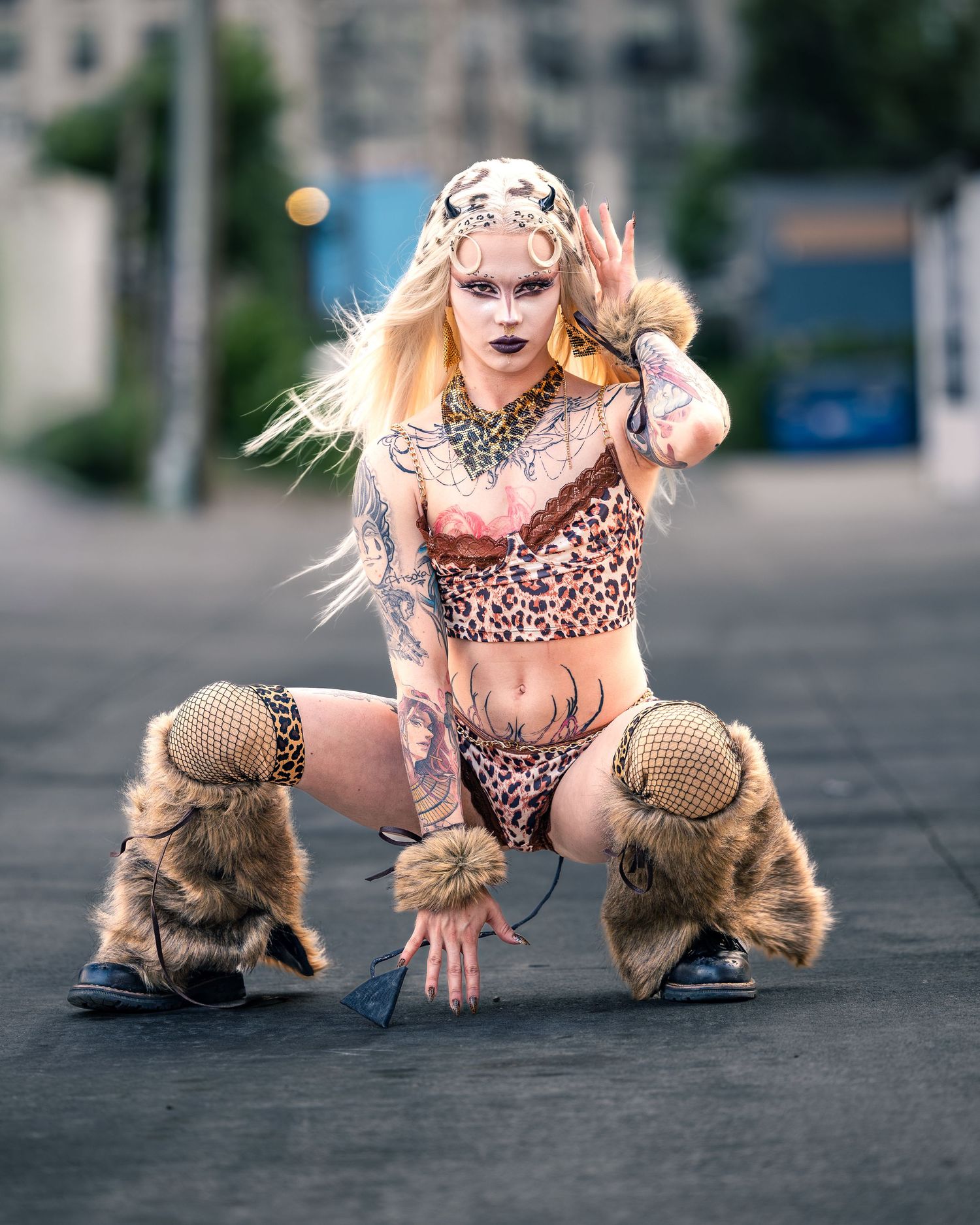 Tattooed performer in leopard print outfit with fur boots and cheetah mask crouches on pavement.