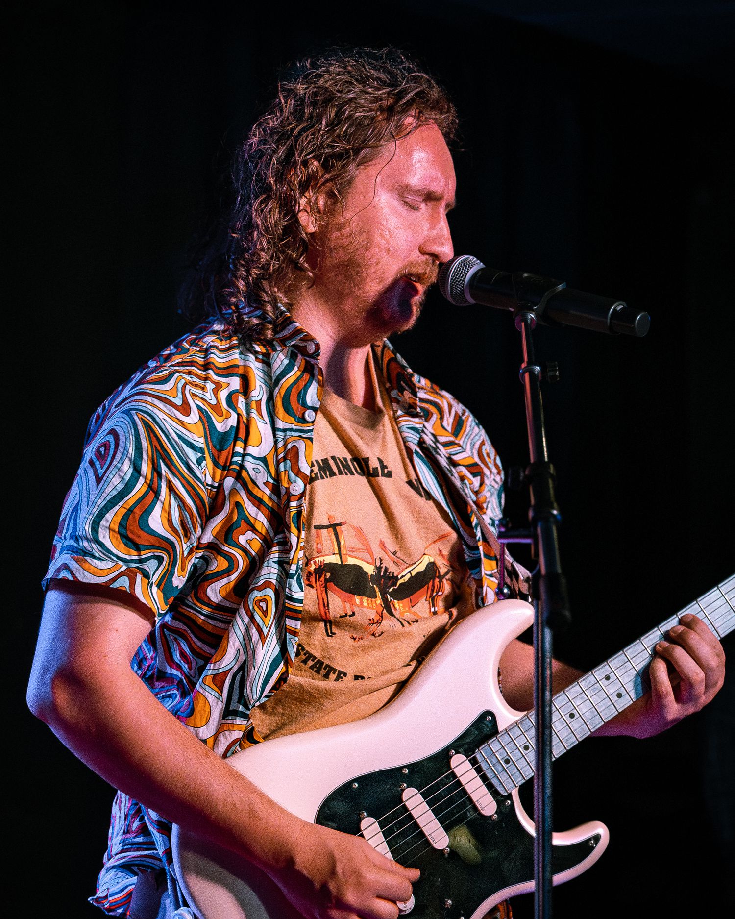 Curly-haired guitarist in colorful patterned shirt plays white Stratocaster guitar at live music show.