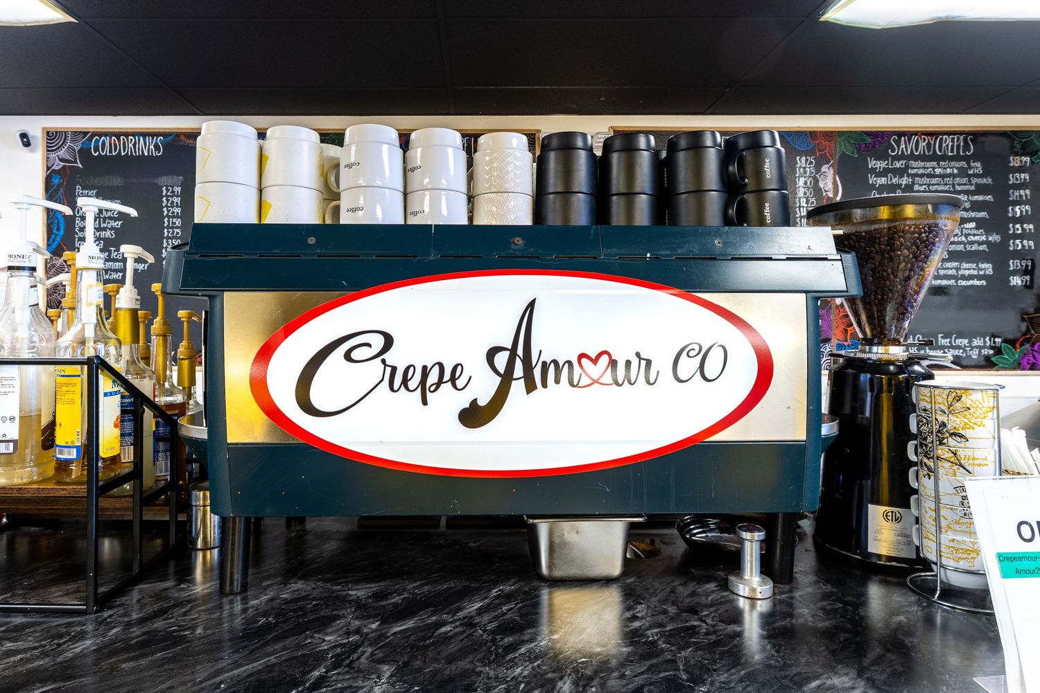 Crepe Amour CO espresso machine at a cafe counter with coffee equipment and menu boards in background.