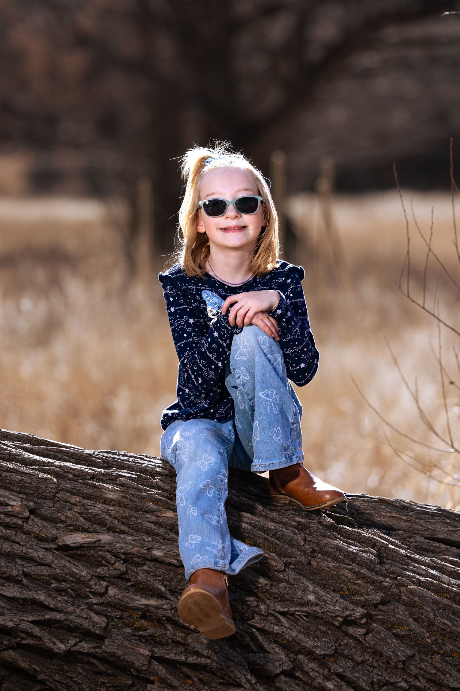 Young girl in sunglasses and jeans sits smiling on a fallen log in an autumn field.