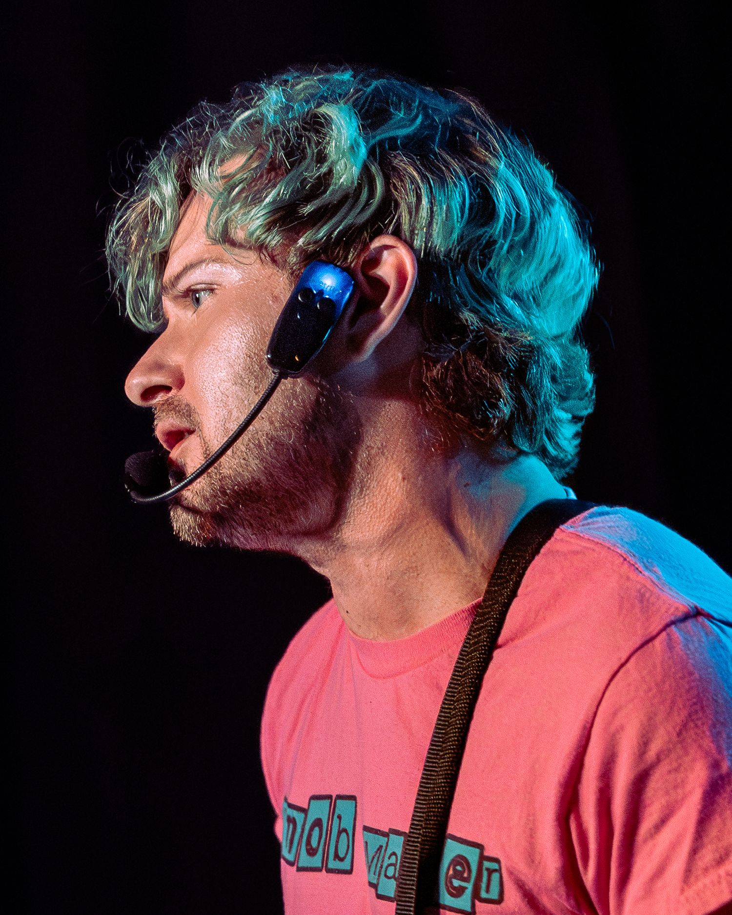 Young man in pink t-shirt wearing headset mic and earpiece performing on stage under teal lighting.