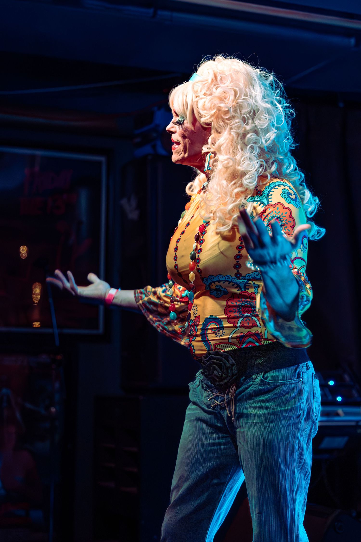 Performer in colorful blouse and big blonde wig gestures on stage under blue and red lights.