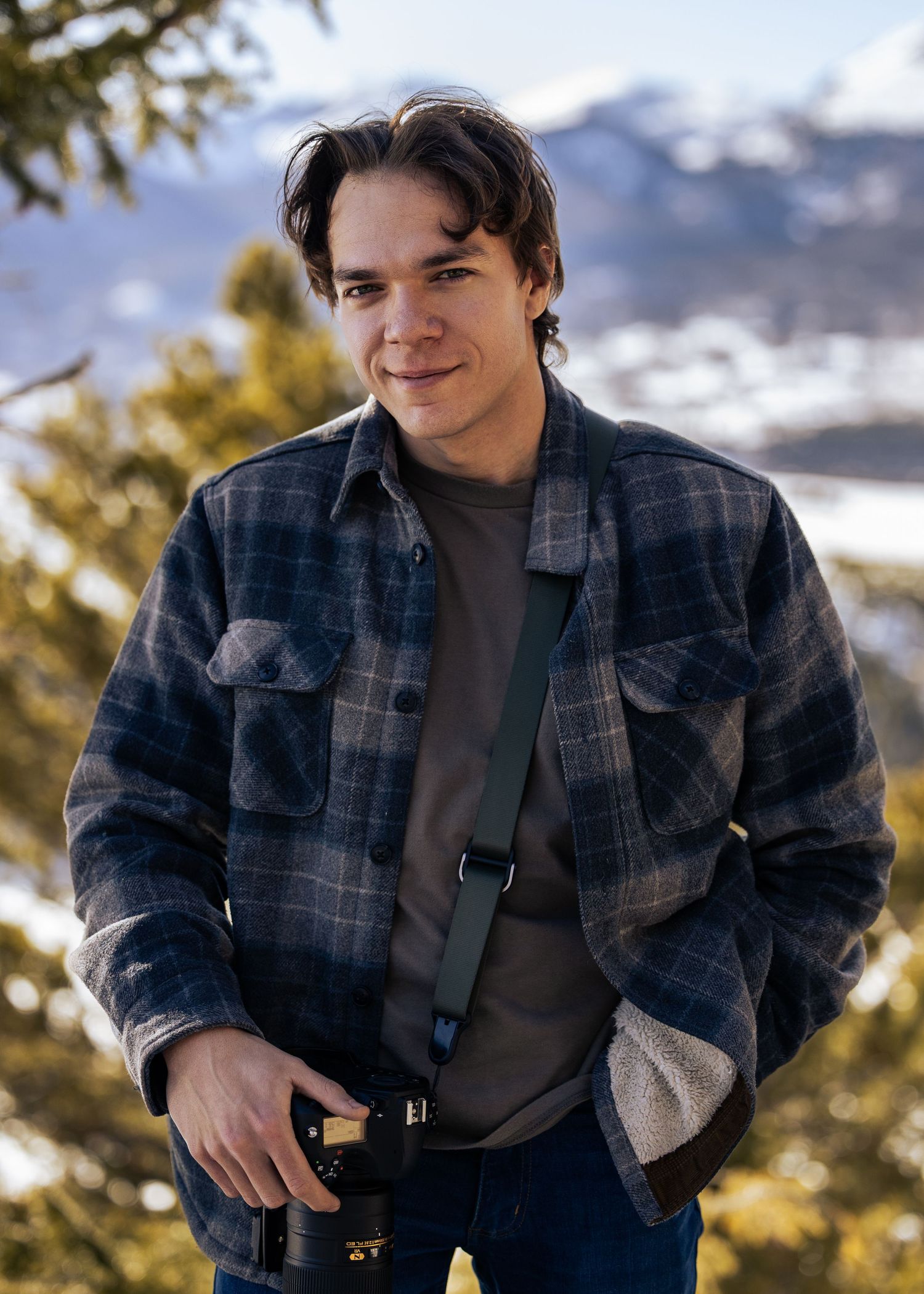 Photographer in plaid jacket and camera strap against snowy mountain backdrop.