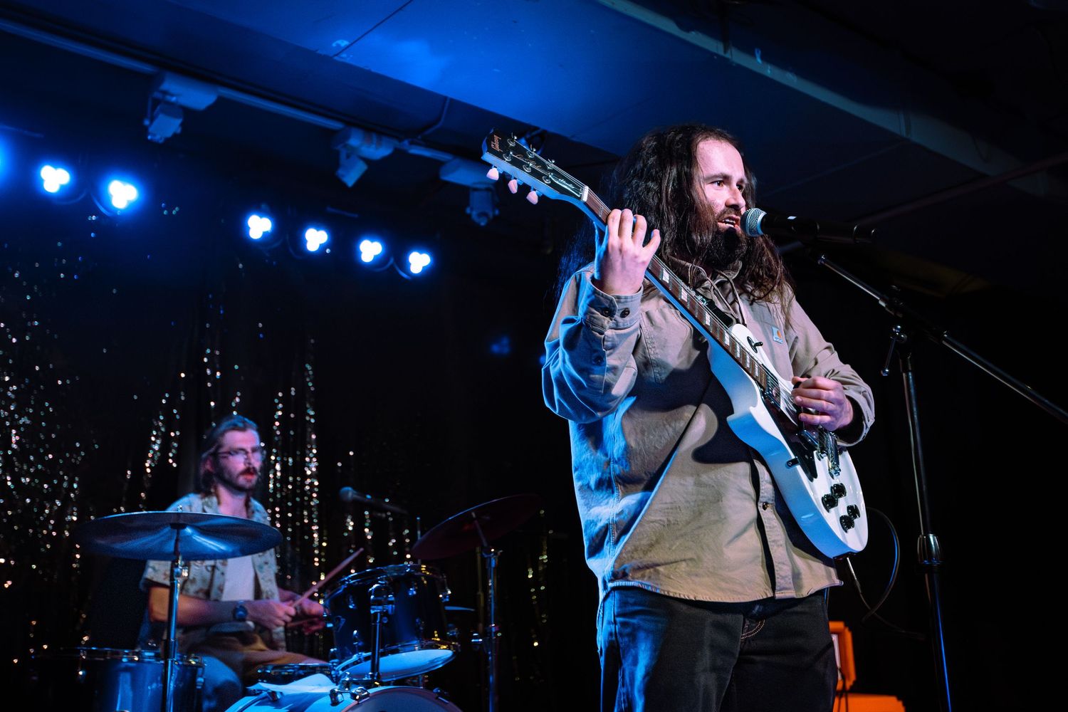 Long-haired guitarist performing on stage with drummer in background under sparkling stage lights.