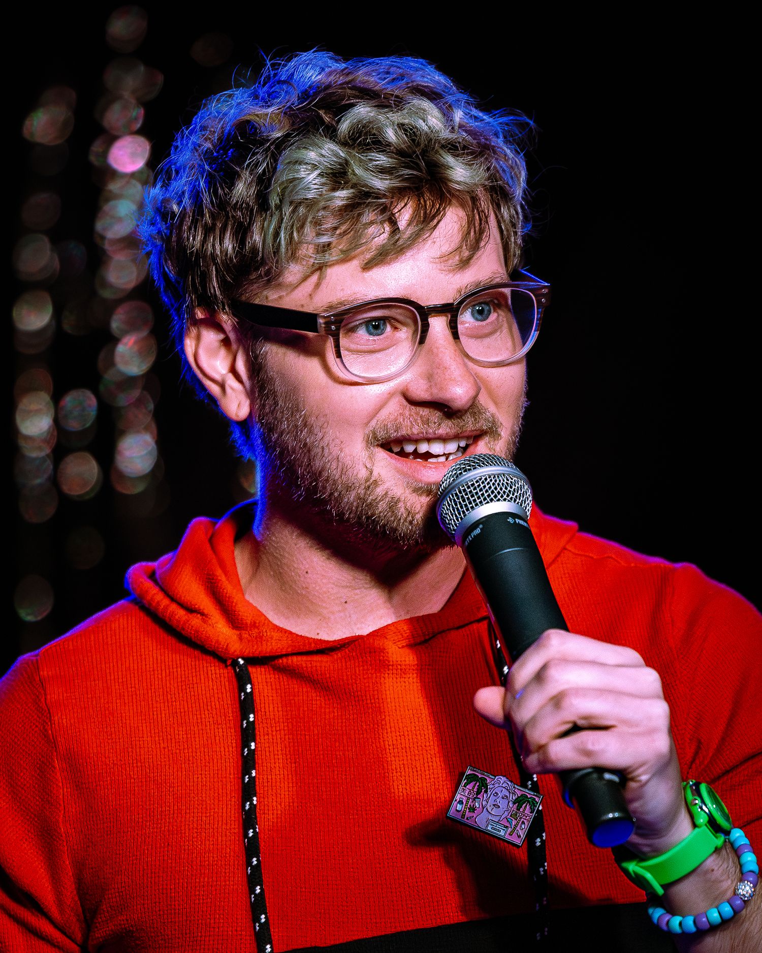 A curly-haired man with glasses and red hoodie smiling while holding a microphone on stage.