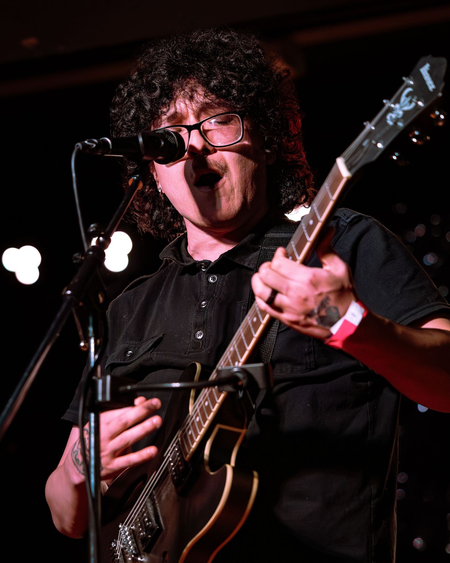 Curly-haired guitarist in black shirt plays electric guitar at microphone on stage.