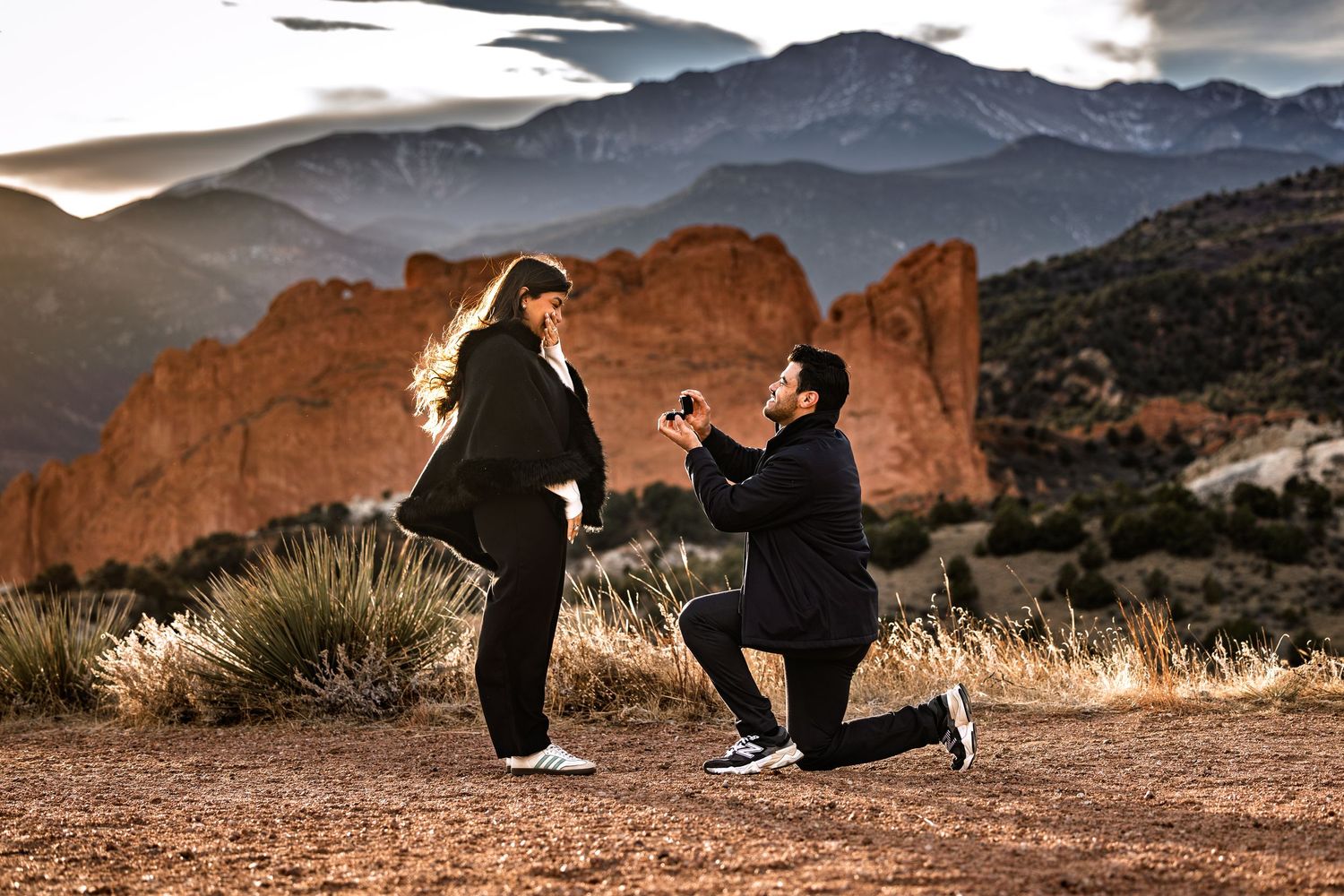 A silhouetted couple shares a romantic proposal moment against dramatic red rock formations at sunset.