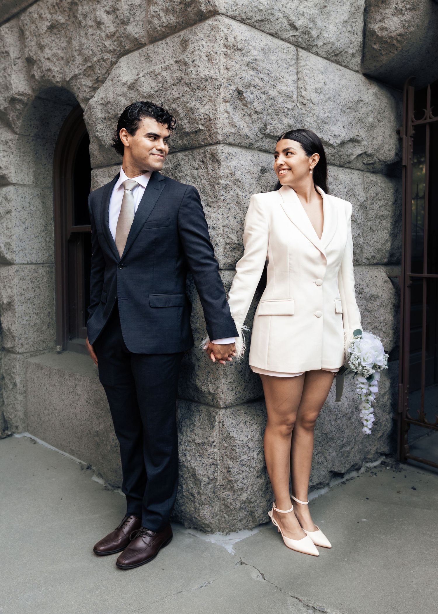 Couple holding hands in wedding attire, man in dark suit, woman in white blazer dress, posing by stone building.