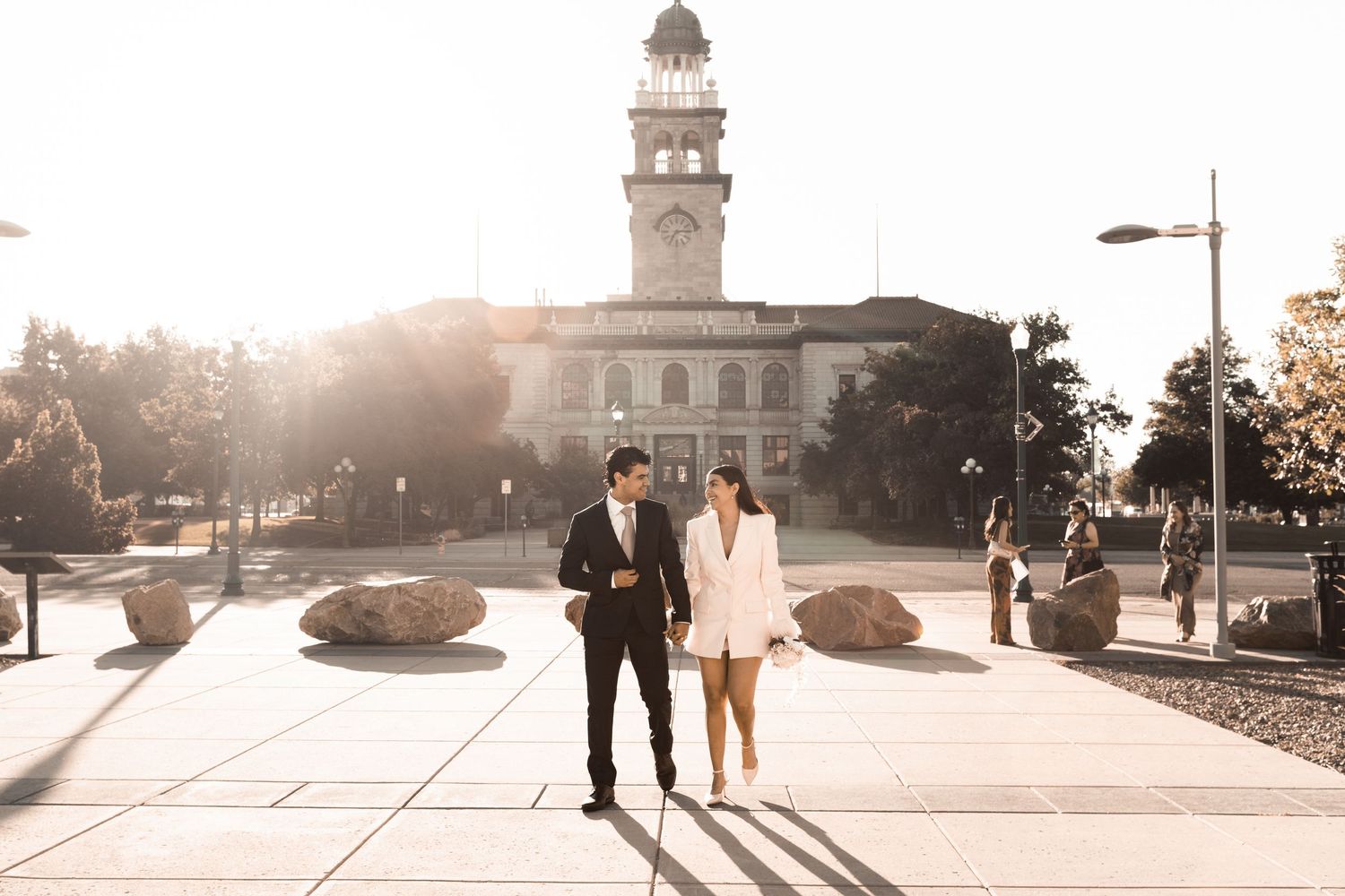 A couple in formal attire walks hand in hand near a historic clock tower building at sunset.