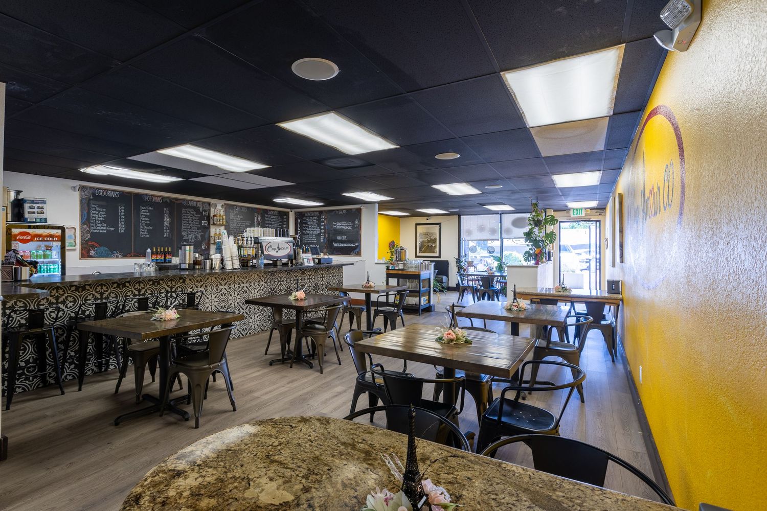 Cozy cafe interior with black ceiling, yellow accent wall, wooden tables, and chalkboard menu.