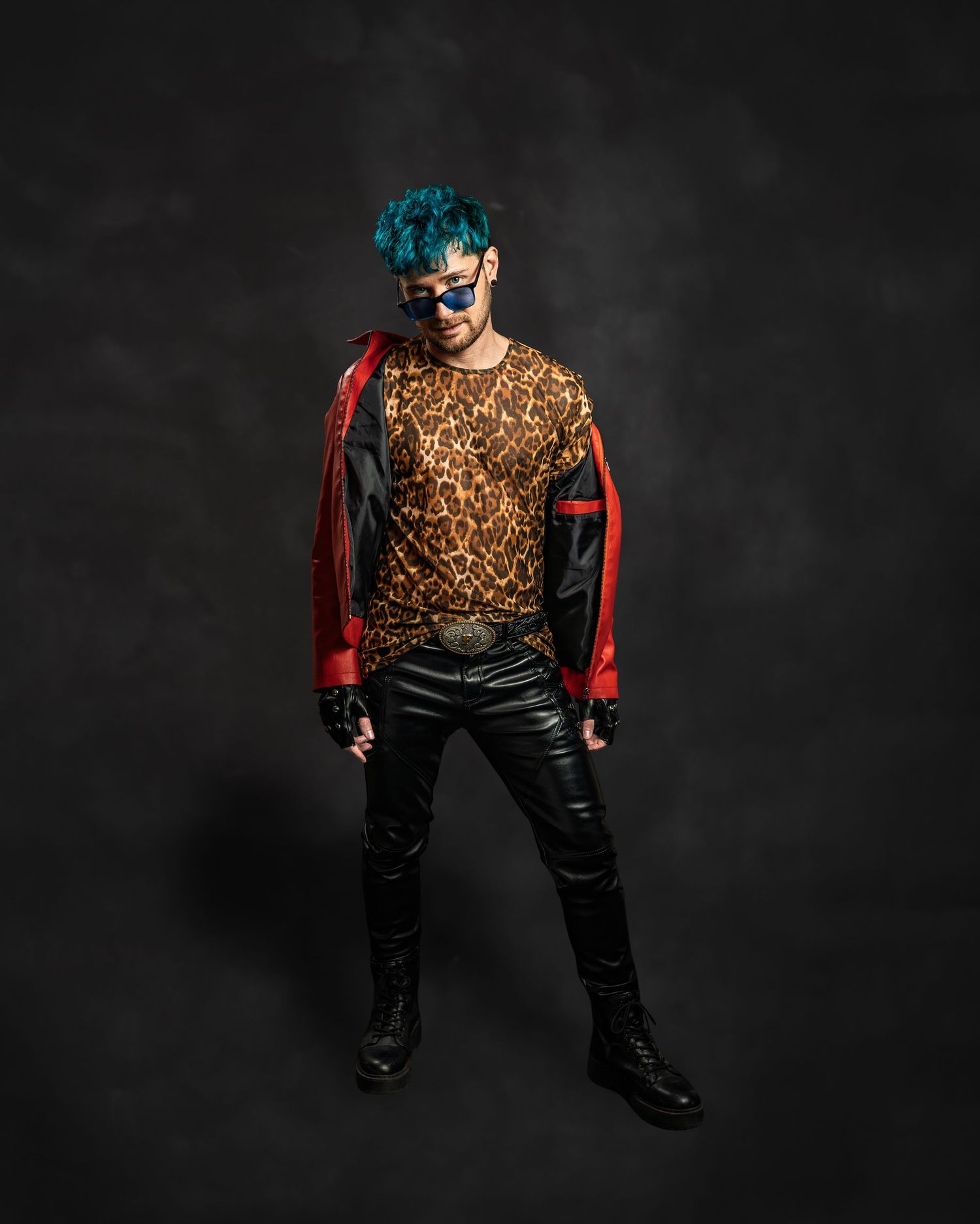 Man with teal hair in leopard print shirt, red leather jacket, and black boots posing confidently.