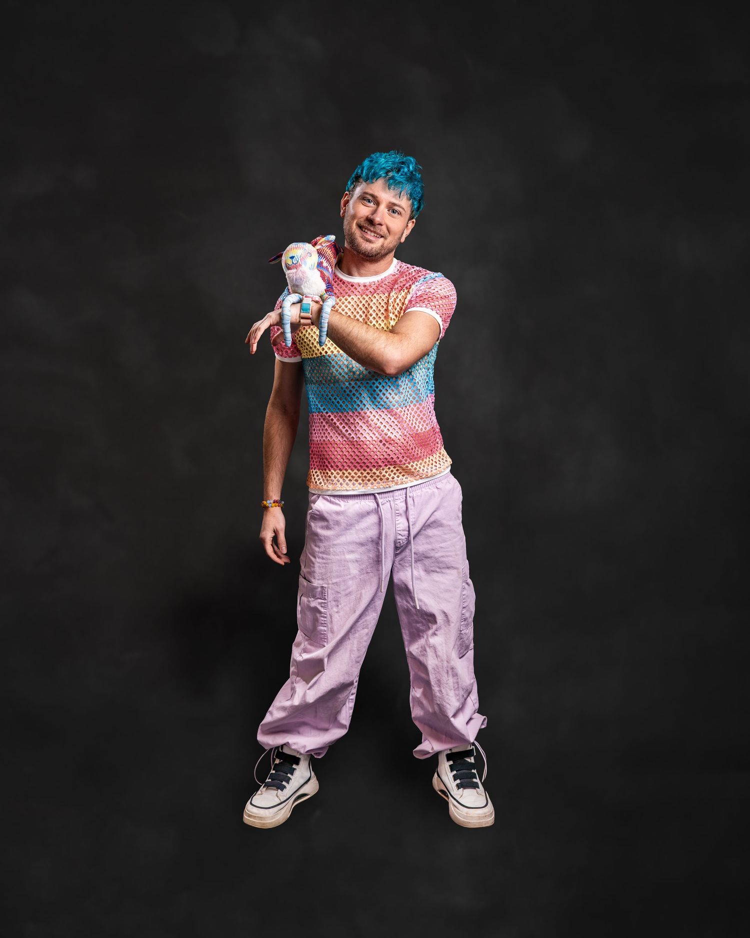 Person with teal hair wearing colorful striped crop top and lavender cargo pants holding a plush toy against dark background.