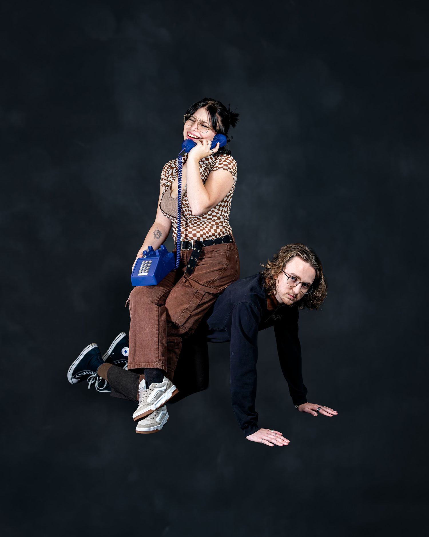 Two friends pose playfully in a studio, one sitting on the other's back while talking on a blue retro phone.