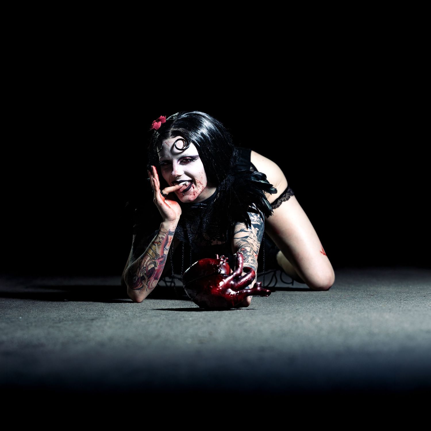 Gothic woman in dark makeup with tattoos poses dramatically on floor with red apple in dark studio setting.