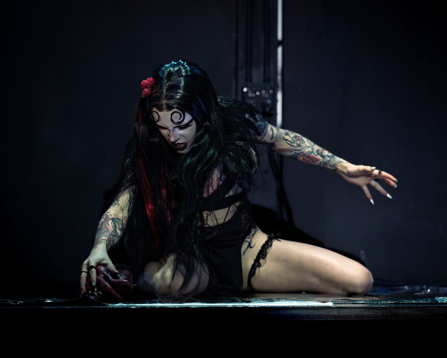 Gothic performer with horror makeup and tattoos crawls dramatically across a dark wet stage floor.