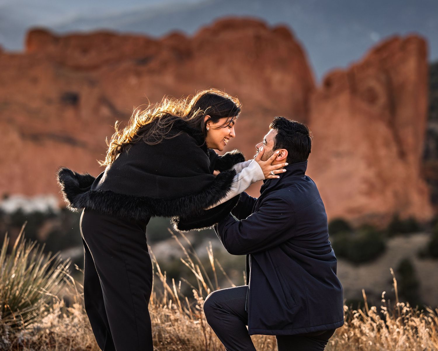 A couple in formal attire embraces in front of red rock formations during a romantic photoshoot at sunset.
