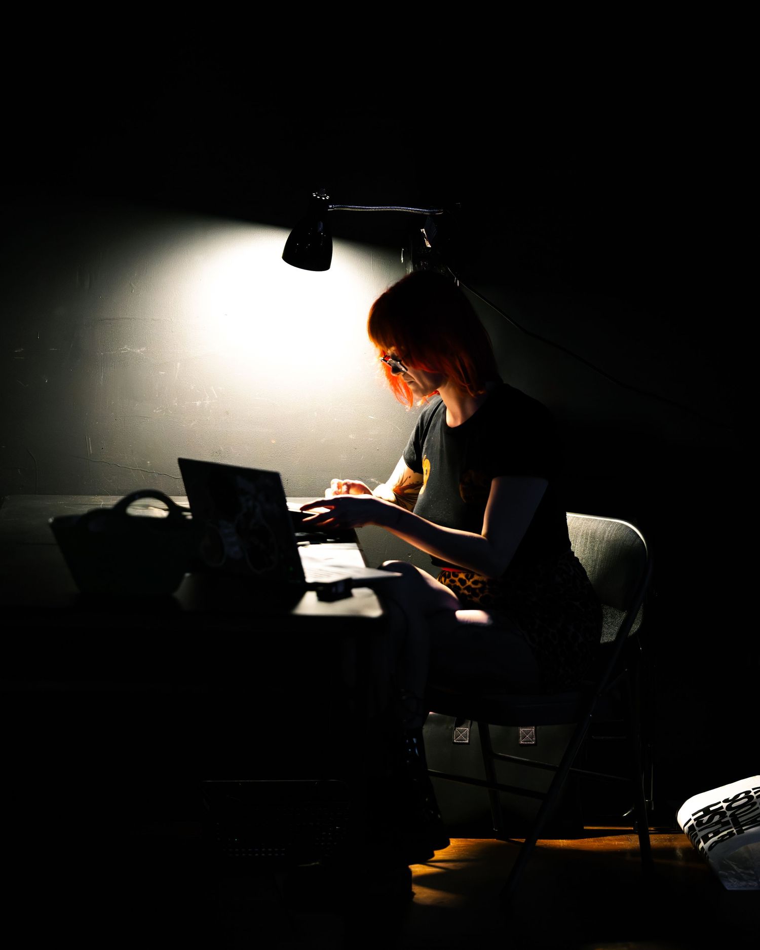 Person working on laptop under desk lamp in dark room, dramatic lighting highlighting focused work session.