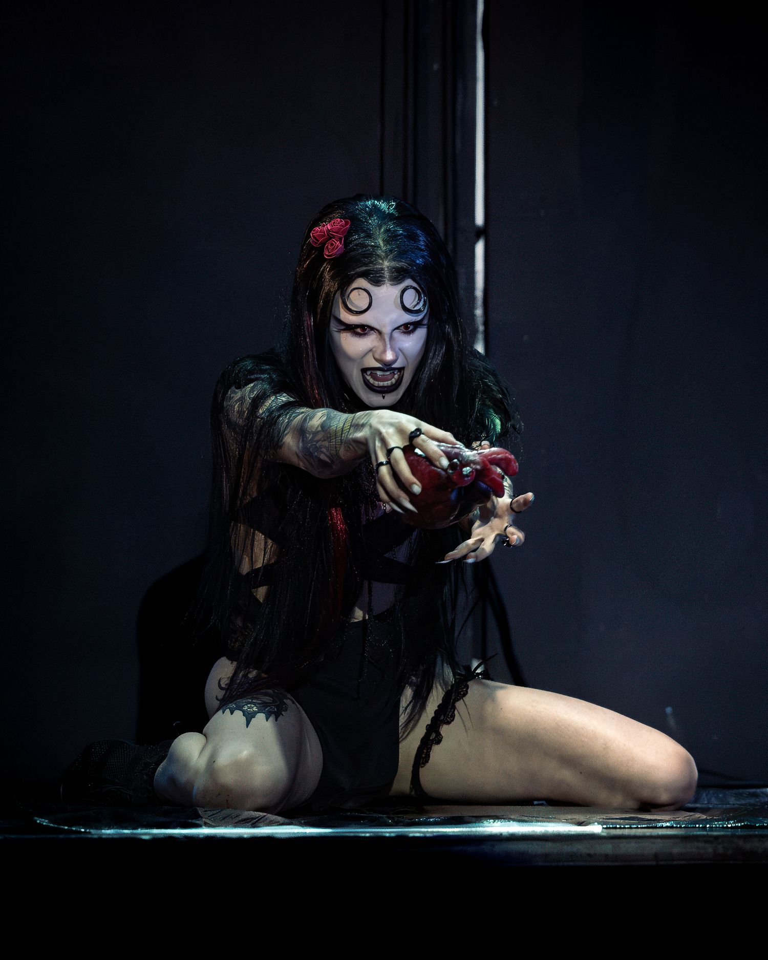 Gothic performer in horror makeup and tattered black costume kneels on stage holding a bloody prop.