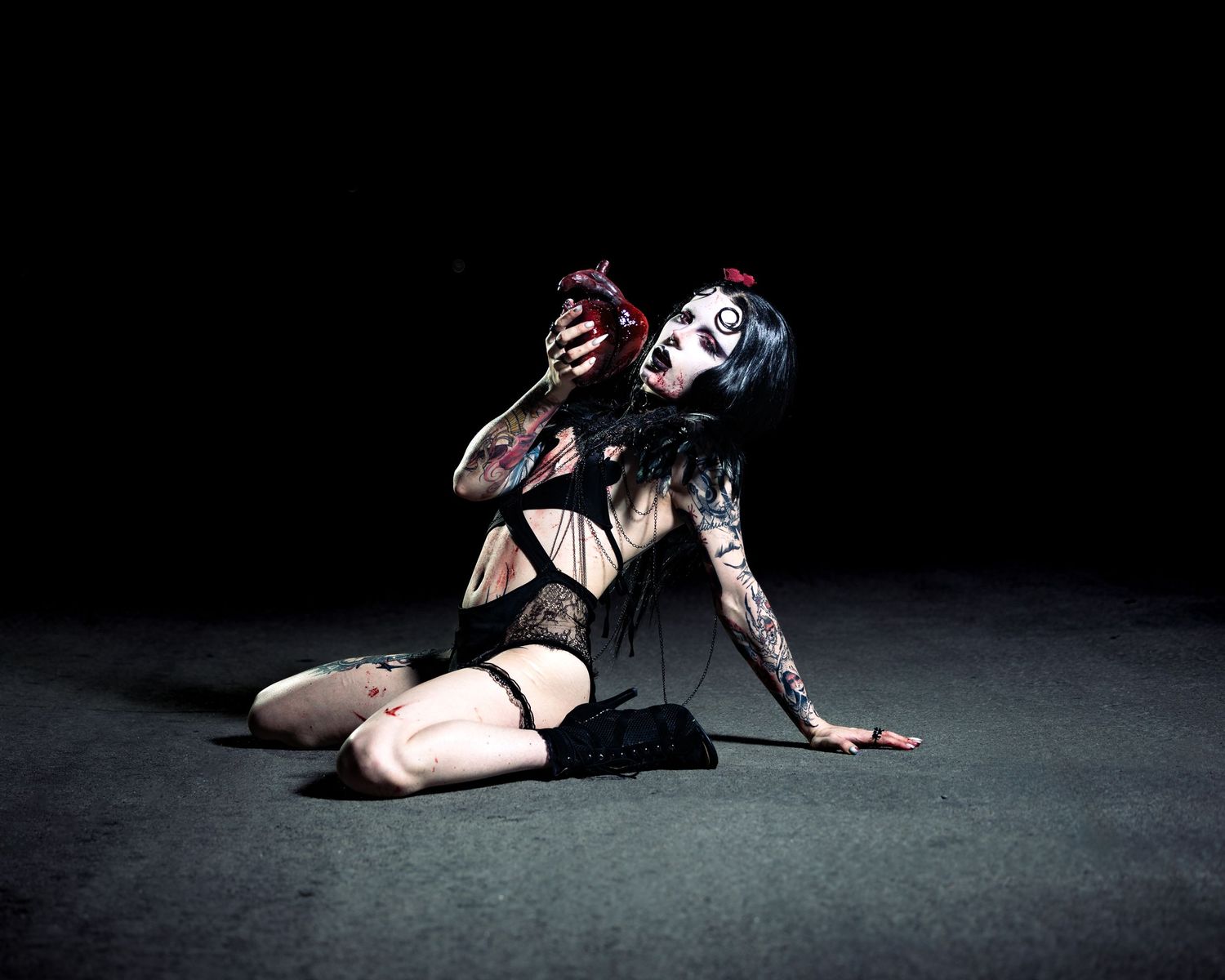 Tattooed performer in gothic makeup and black lingerie kneeling dramatically on floor holding red object.