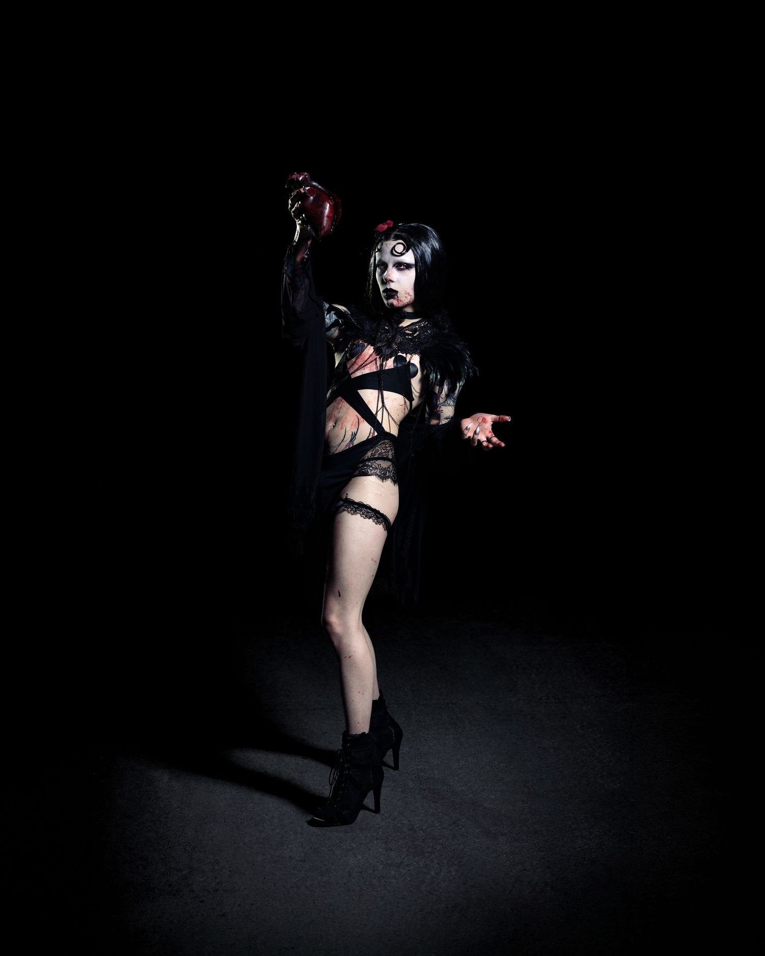 Dark gothic performer in black costume and dramatic makeup dancing in high heels against black background.