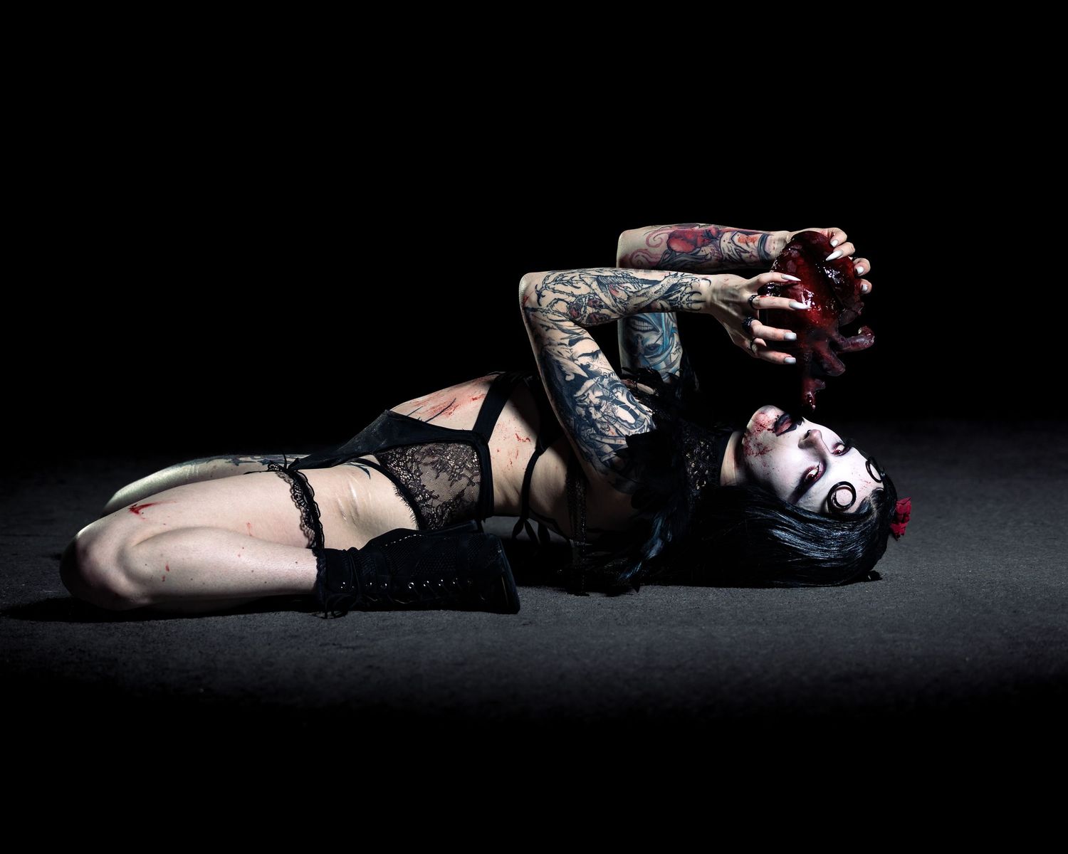 Tattooed woman in black lingerie lying on dark floor in dramatic gothic pose with roses.
