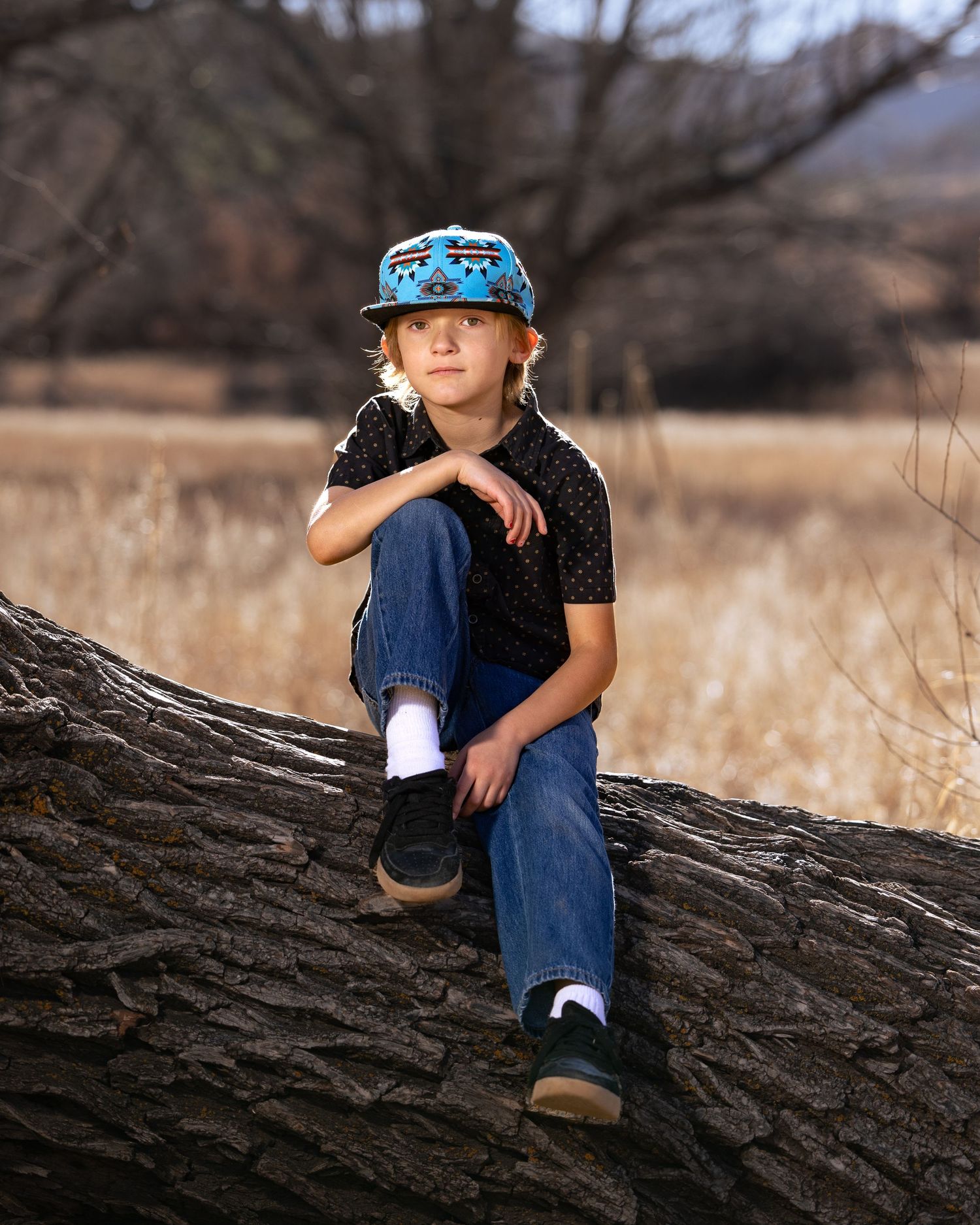 A young boy in jeans and a blue cap sits confidently on a fallen tree trunk in a golden field.