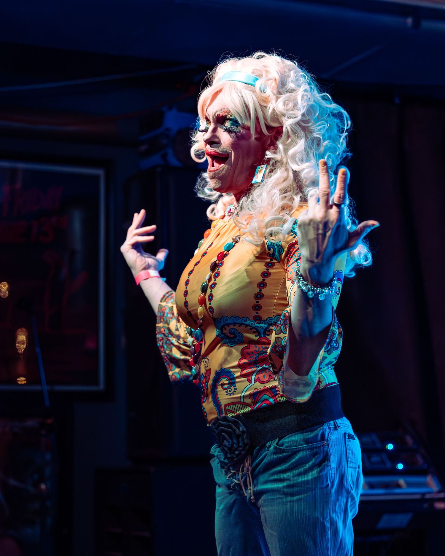 Drag performer in blonde wig and vibrant patterned top gesturing on stage with blue lighting.
