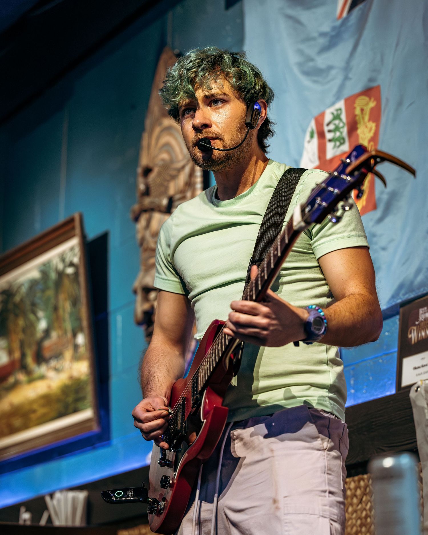 A musician performs on stage with an electric guitar while wearing a light colored t-shirt against a blue wall background.