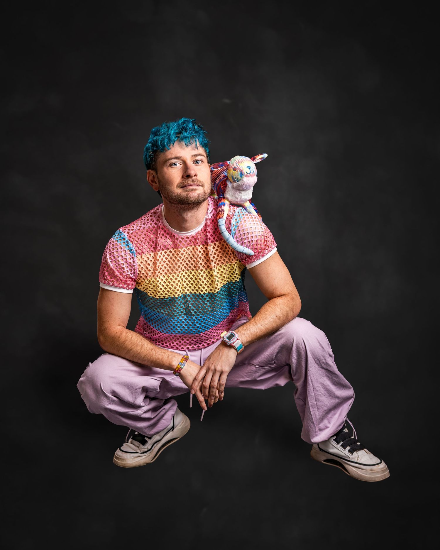 Person with blue hair wearing rainbow mesh shirt and lavender pants, crouching with a small unicorn toy on shoulder.