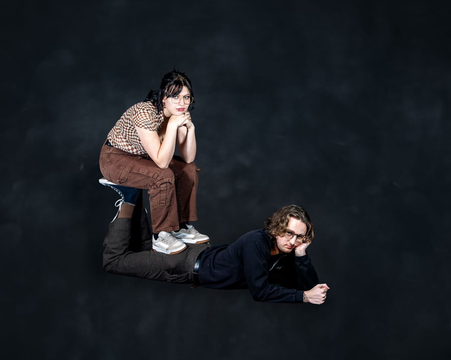 Two young people posing against dark background, one kneeling and one lying down, in casual stylish outfits.