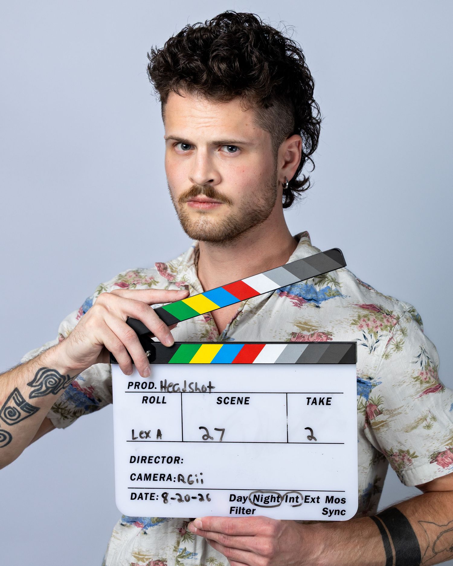 Young man with curly hair and tattoos holds a film clapperboard on a white studio background.