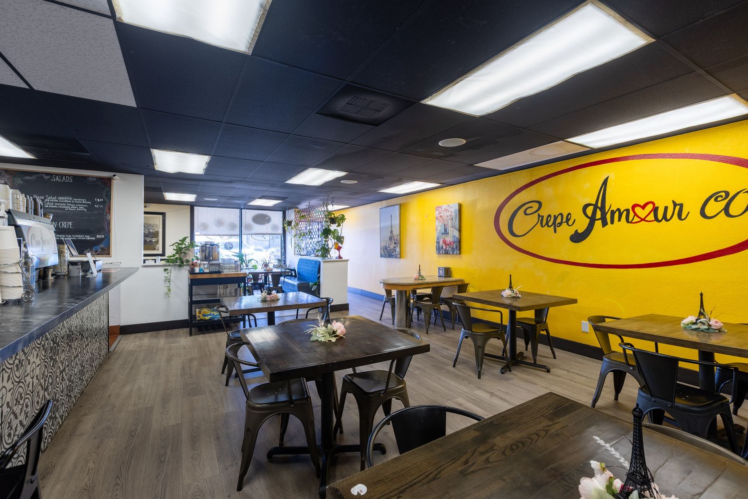 Interior of Crepe Amour cafe with wooden tables, chairs, and a bright yellow accent wall with logo.