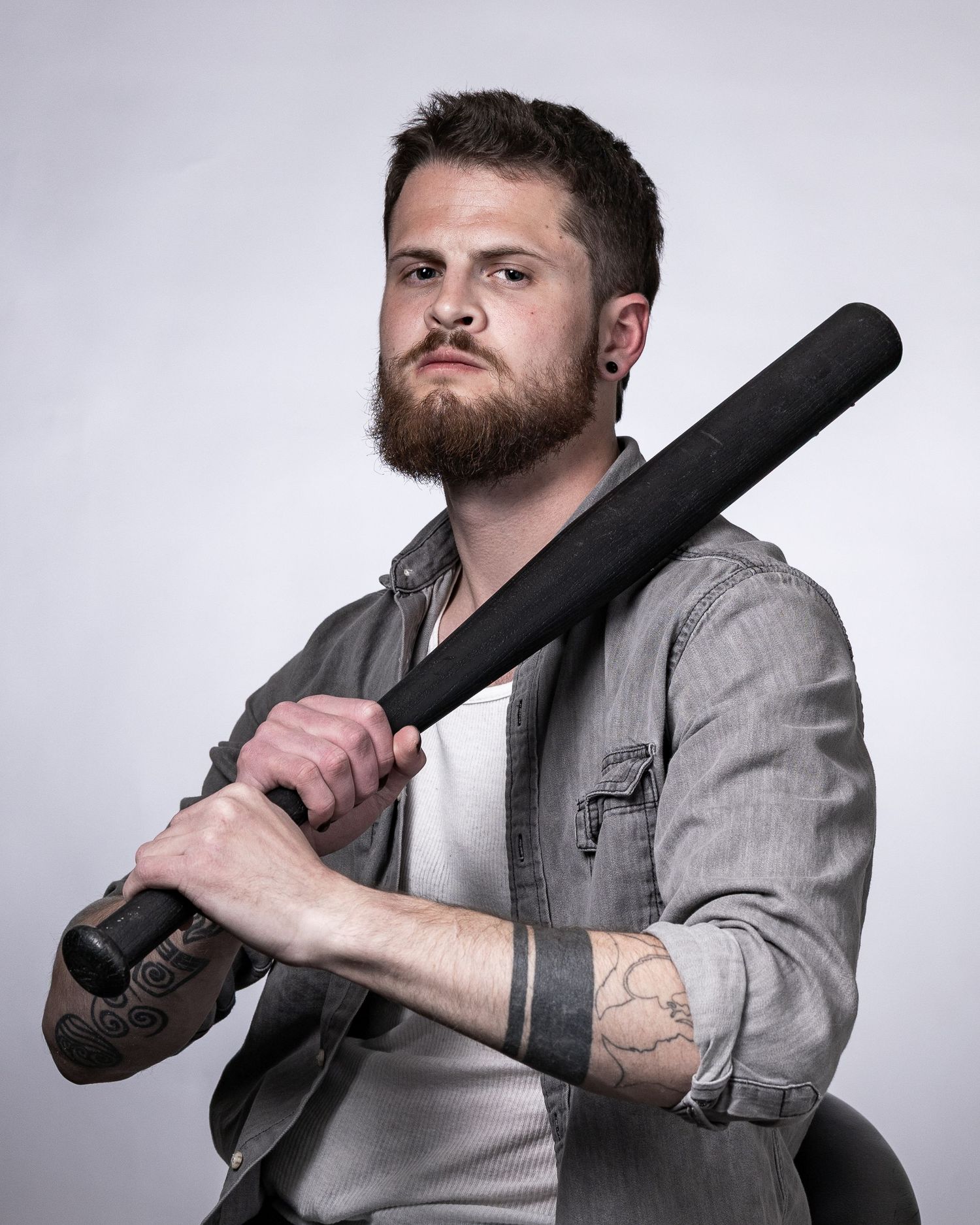 Bearded tattooed man in denim shirt holding black baseball bat over shoulder against gray background.