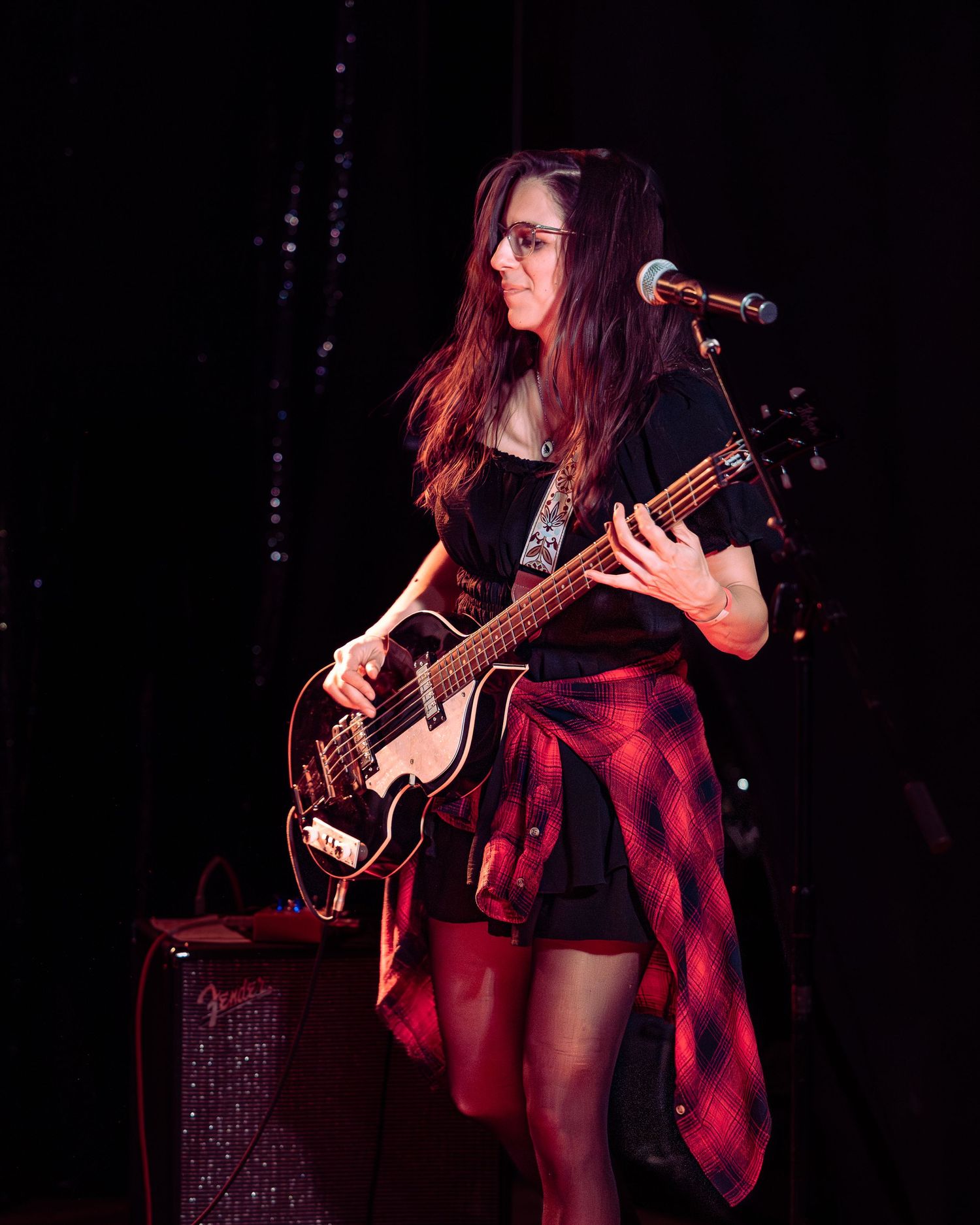 Woman with glasses playing black bass guitar near microphone stand under red stage lighting.