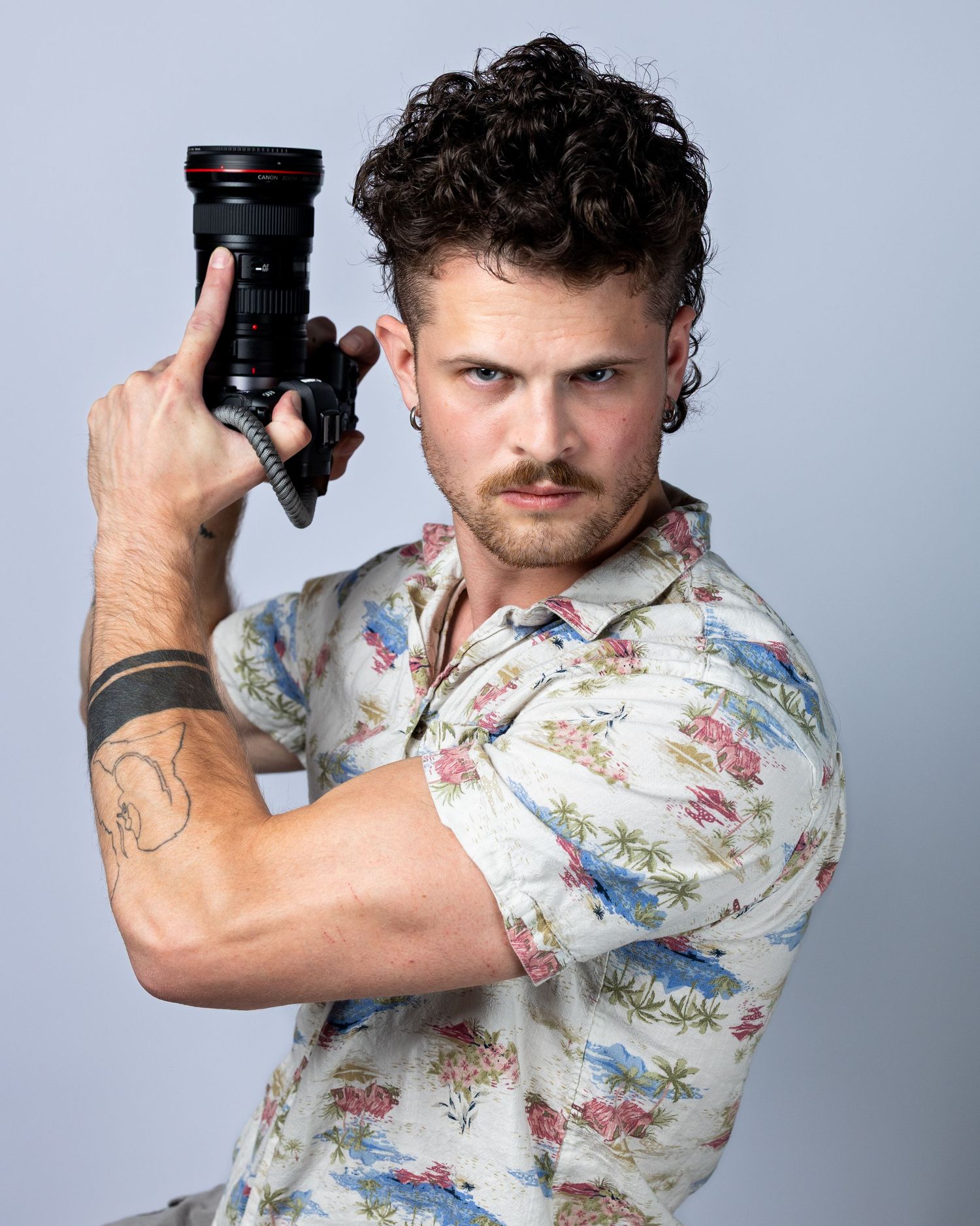Man with curly hair holding a Canon camera in a floral shirt, with a tattoo on his arm, posing confidently.
