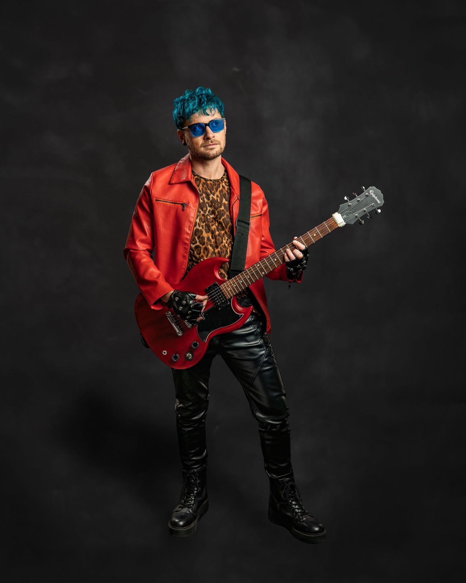 Rock musician with blue hair, red leather jacket, leopard print shirt, and black guitar posing against dark background.