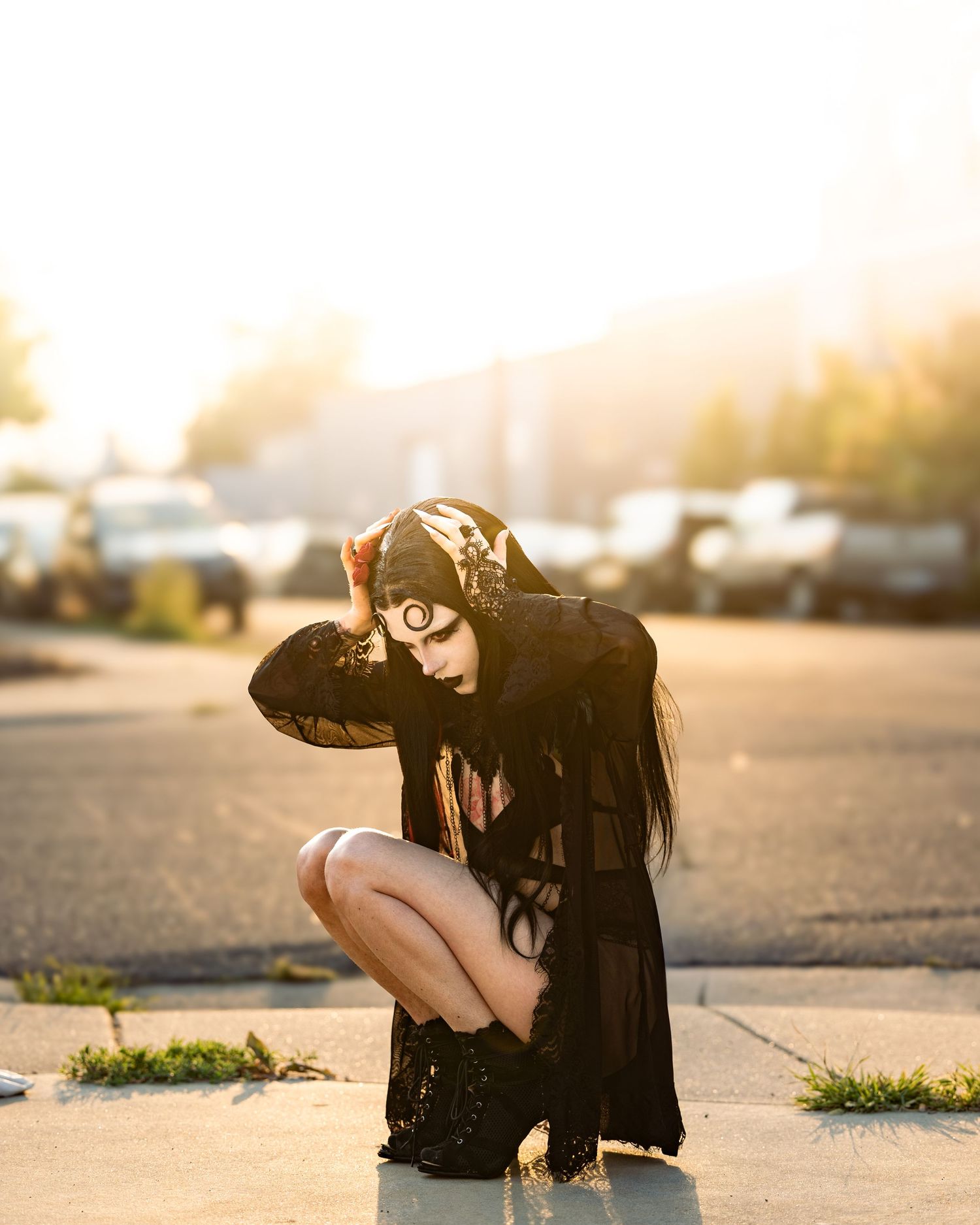 Gothic woman in black outfit with moon makeup crouches on sunlit street in dramatic golden light.