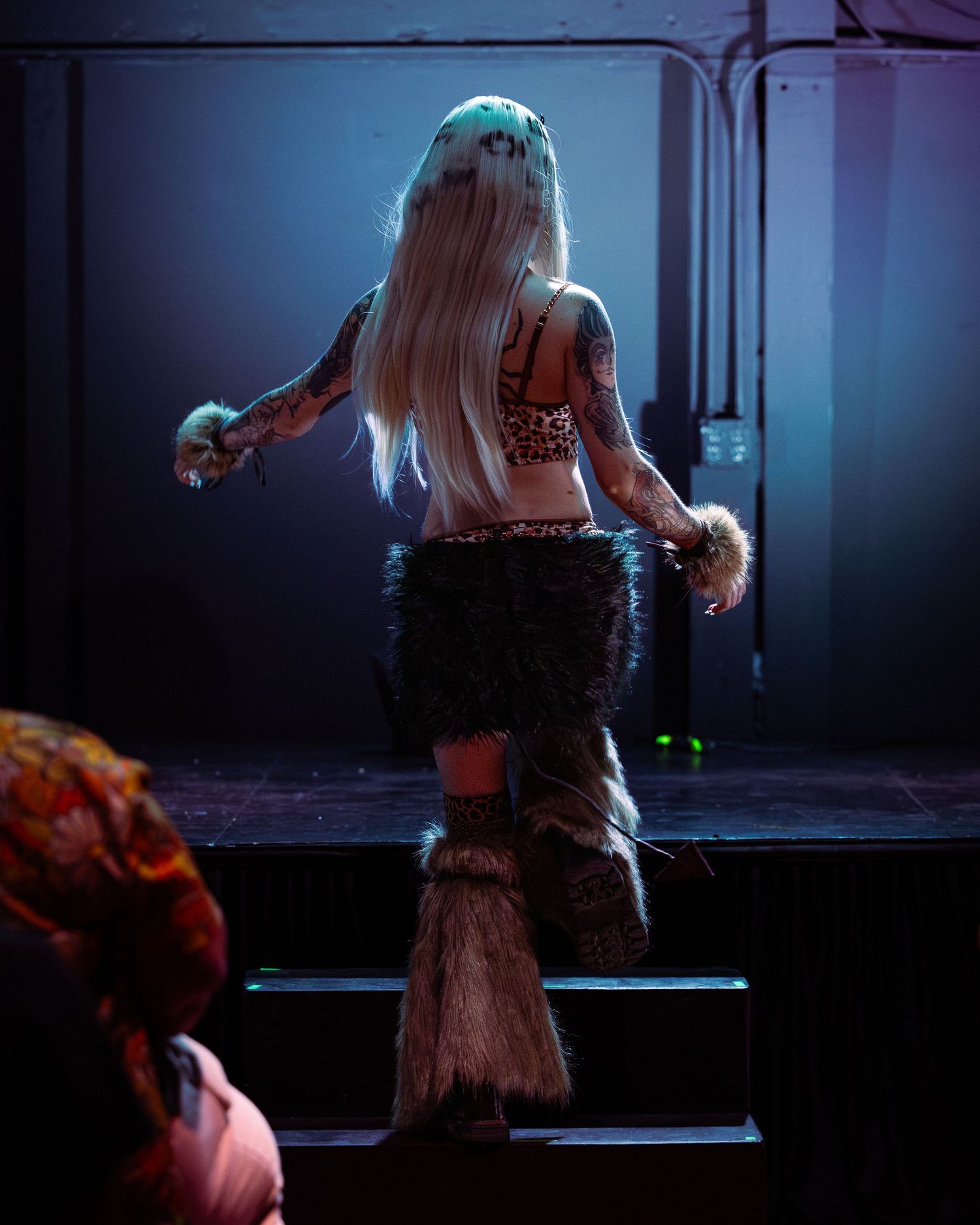 Tattooed performer in fur skirt and crop top with long blonde hair stands on stage under blue lights.