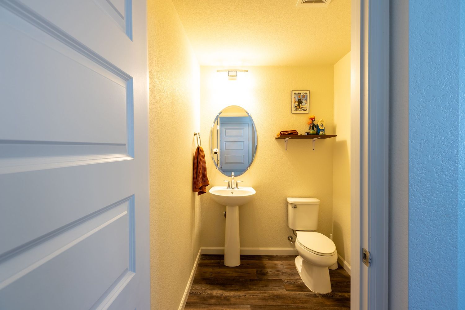 Small bathroom with pedestal sink, oval mirror, white toilet, dark wood floors, and blue door.