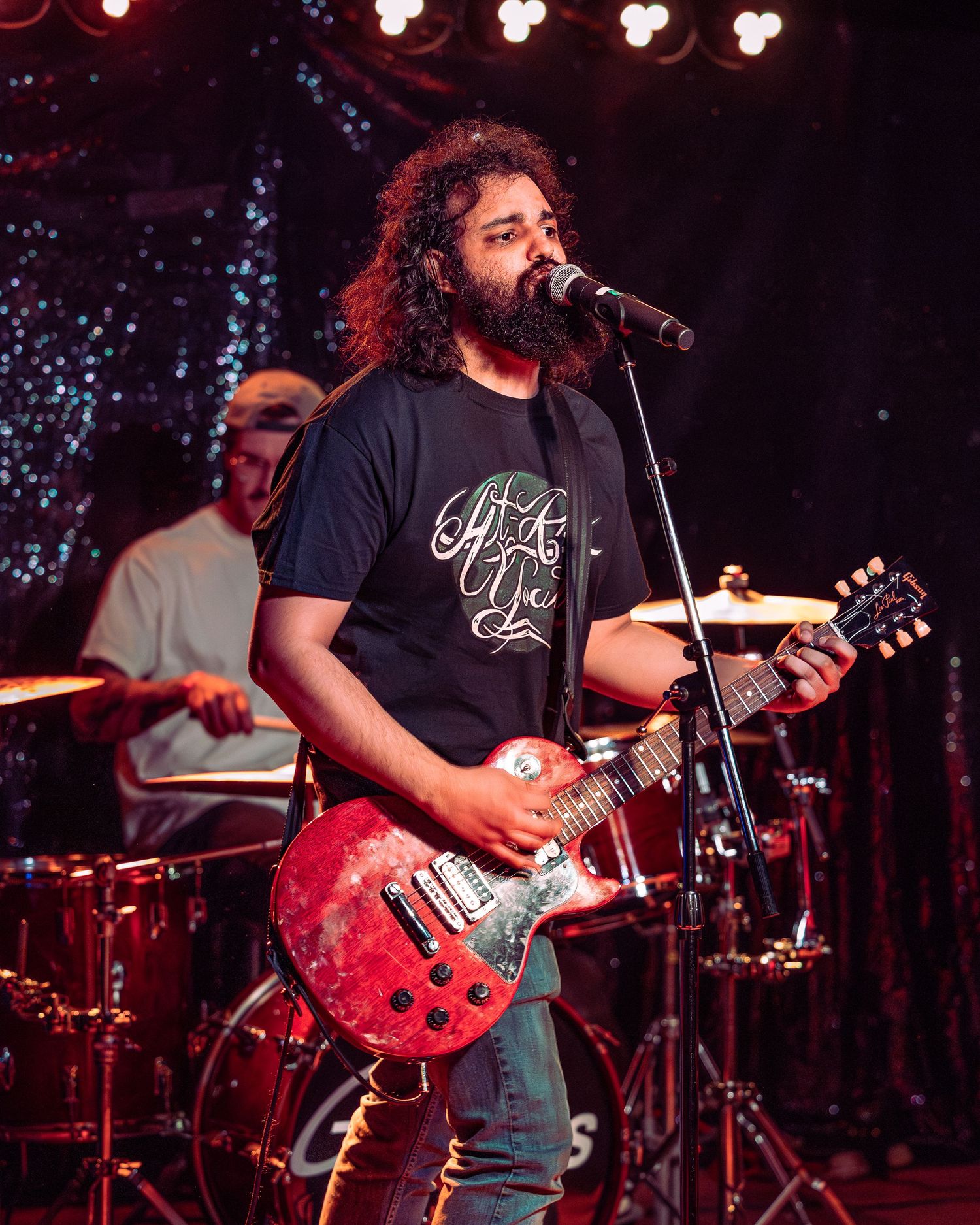 Bearded guitarist with curly hair plays red electric guitar and sings into microphone on stage.