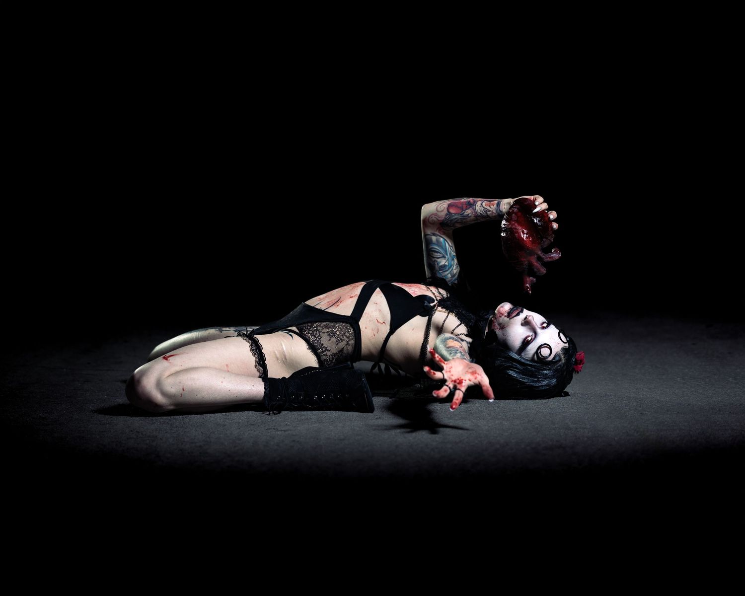 Gothic woman in black lingerie lying dramatically on dark floor with dramatic makeup and tattoos.