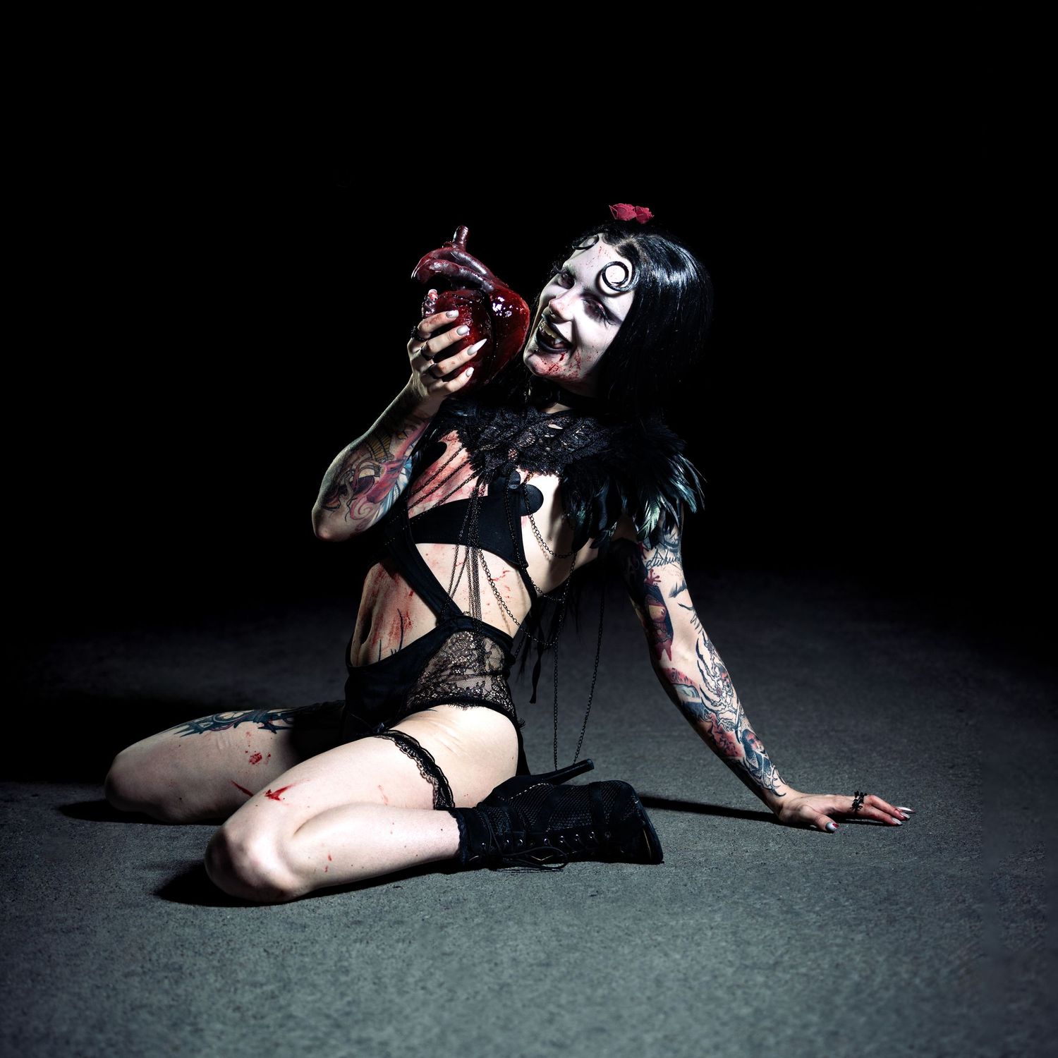 Goth model in dark makeup and black lingerie sits on floor holding red heart prop with tattoos visible.