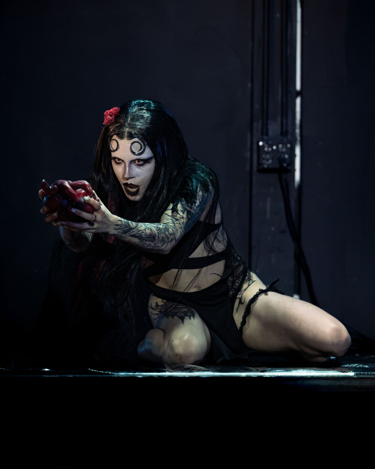 Gothic performer in dark makeup and tattoos kneels on stage holding a red object during dramatic theatrical performance.