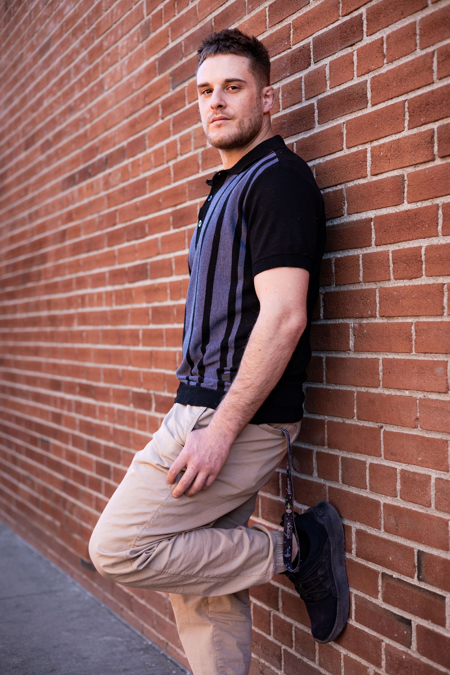 Person in black shirt and khaki pants posing against a red brick wall in various angles.