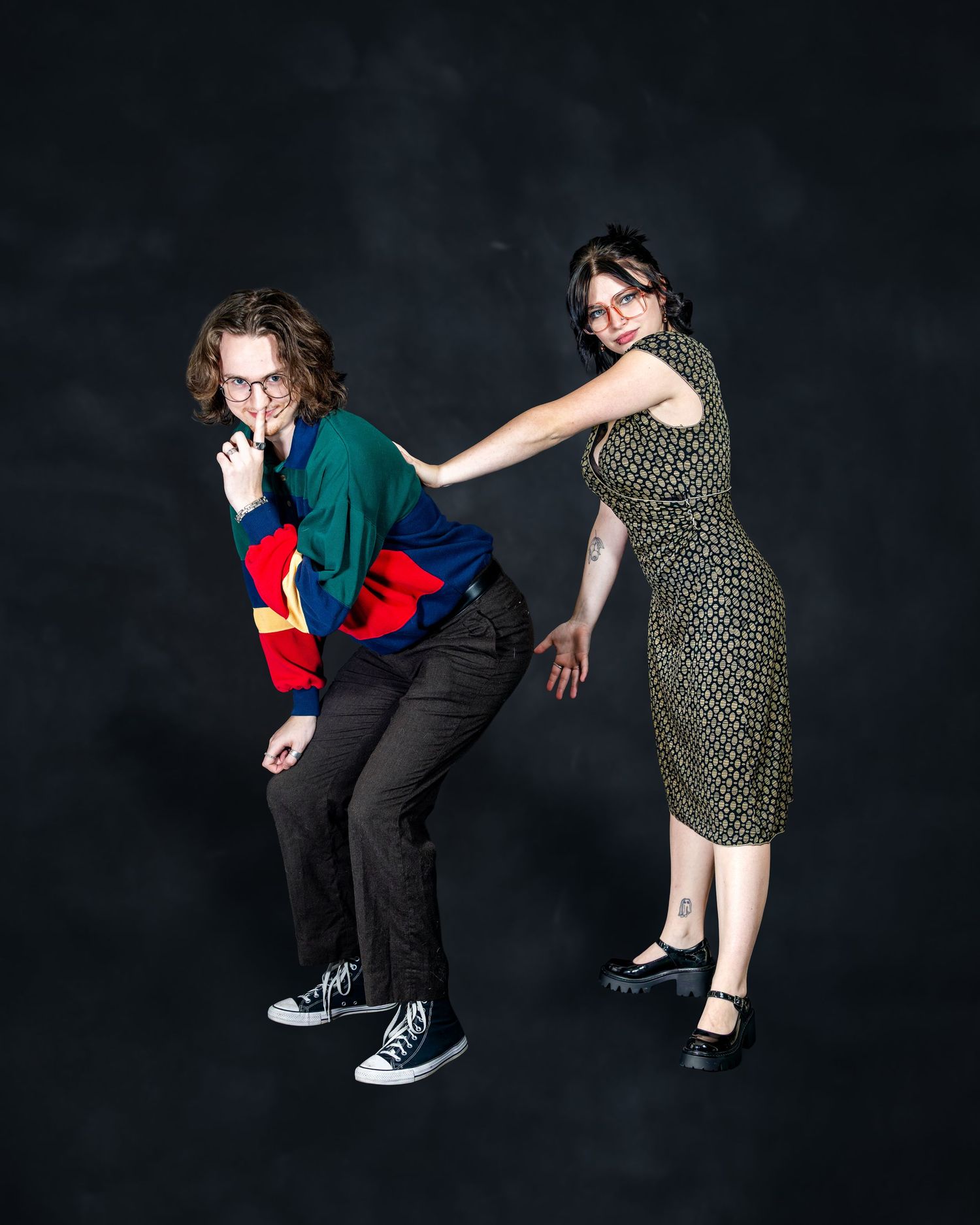 Two young people in colorful outfits strike a fun pose in a dark studio setting.