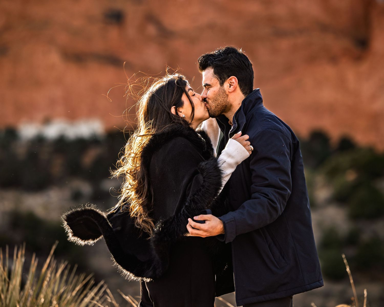 A couple shares a romantic kiss against a backdrop of red rock formations in an outdoor desert landscape.