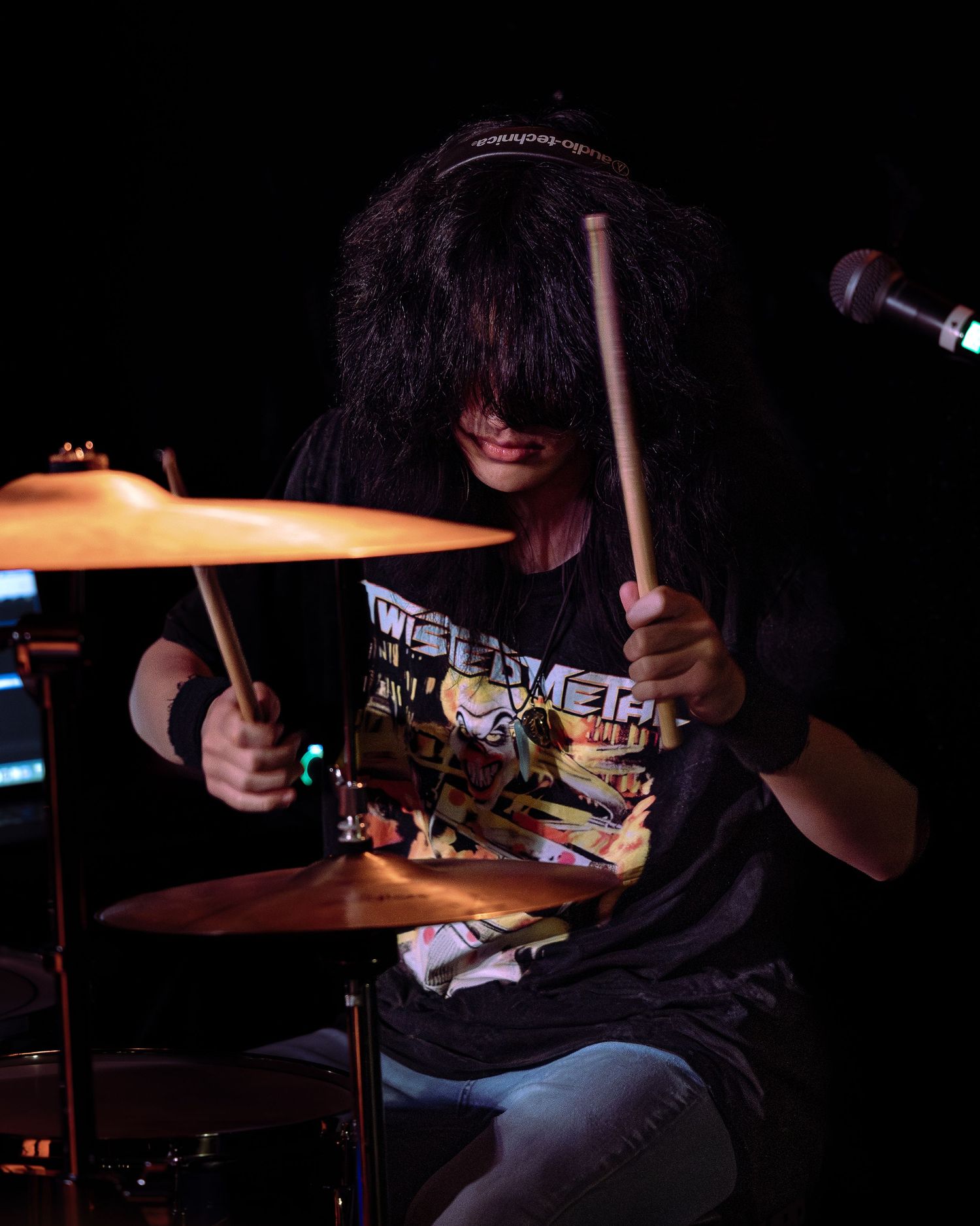 A drummer with long hair plays cymbals on stage, wearing a black graphic tee, holding drumsticks mid-performance.