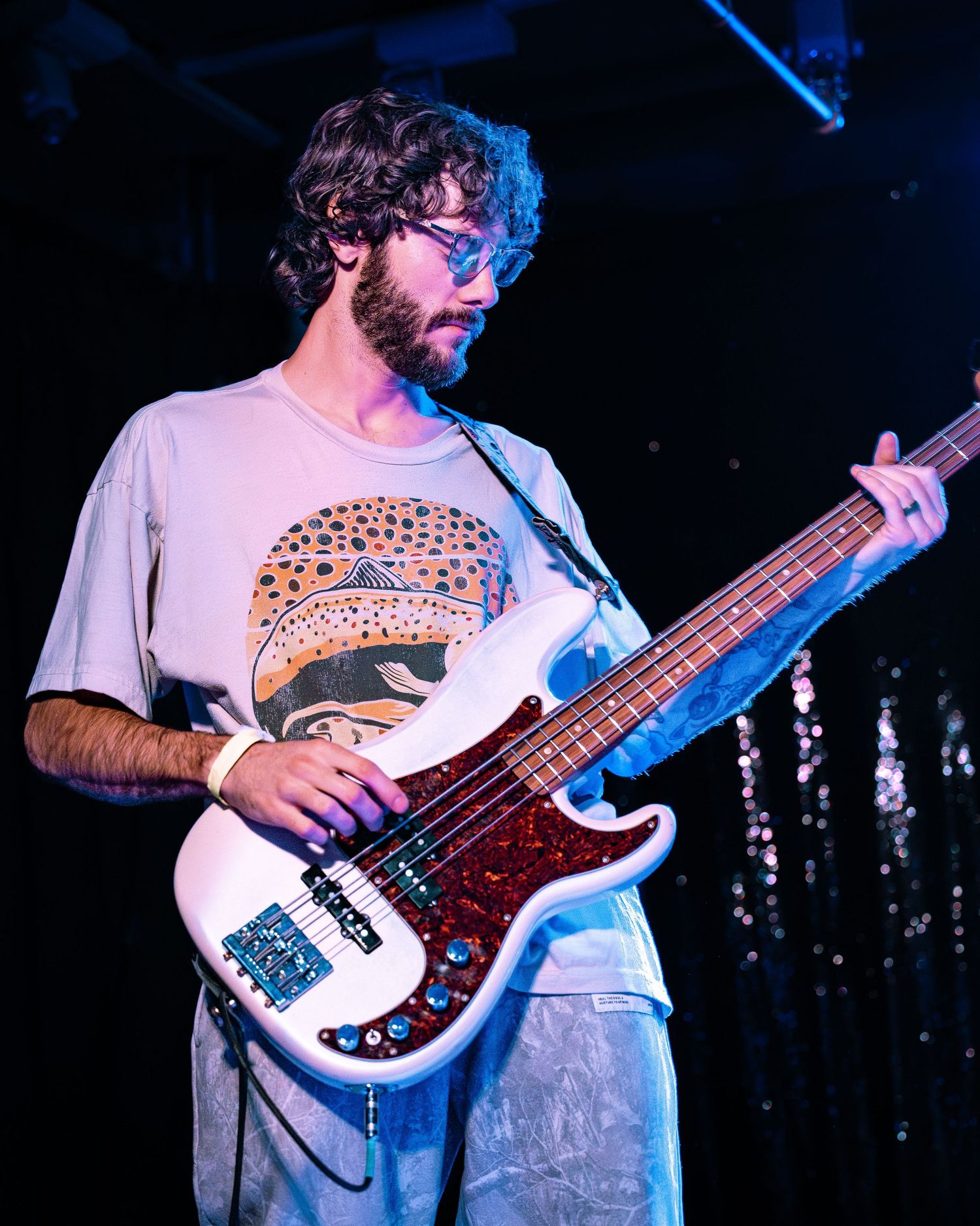 Bearded male bassist in graphic tee plays white electric bass guitar on a dimly lit stage.