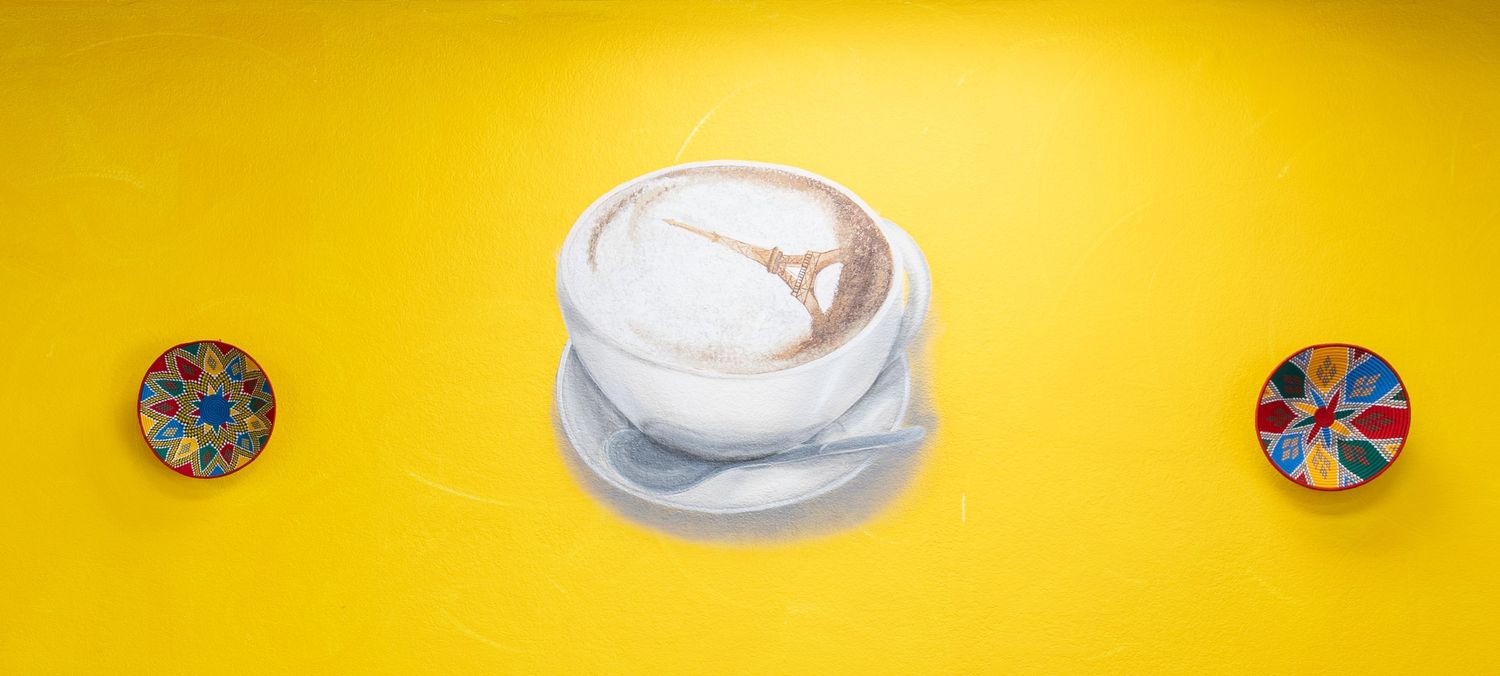 A realistic painting of a cappuccino on a yellow background flanked by two colorful circular objects.